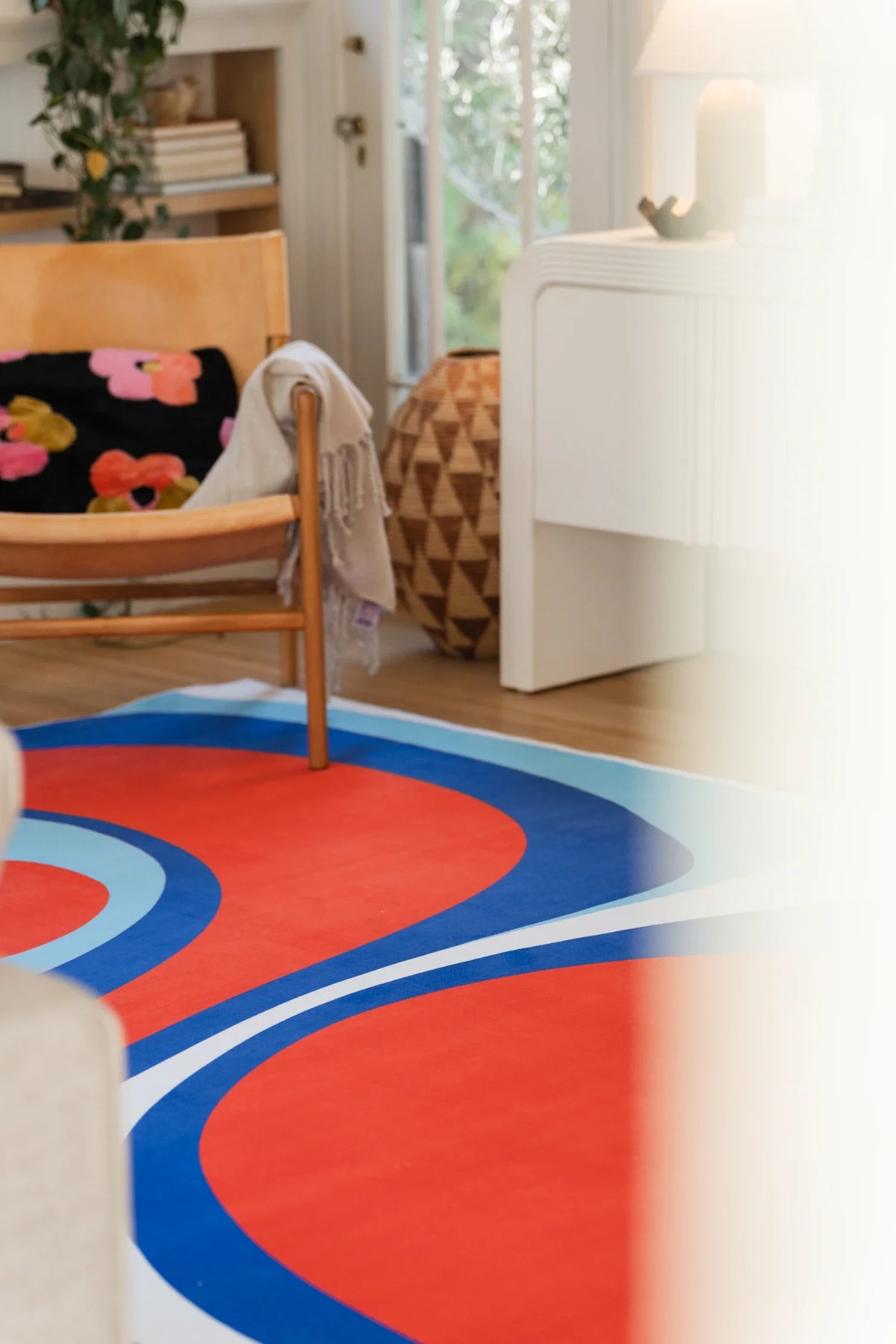 Blue and red Tomato Twist rug in a bright room with a white cabinet, floral pillow, and rounded tomato forms
