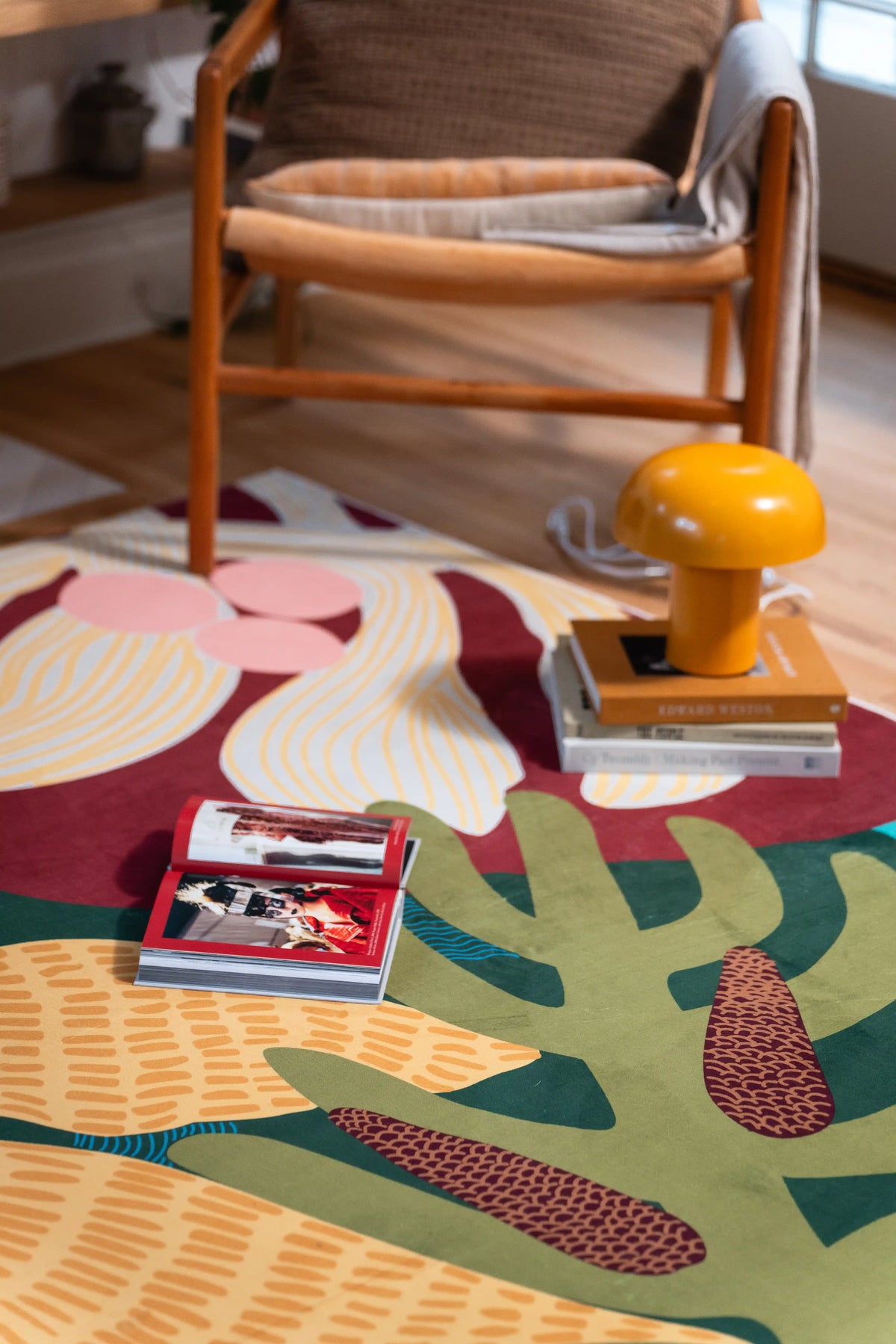 Large cream and pink flowers on green and red sections beneath a wooden armchair, beside a yellow lamp and stacked books.