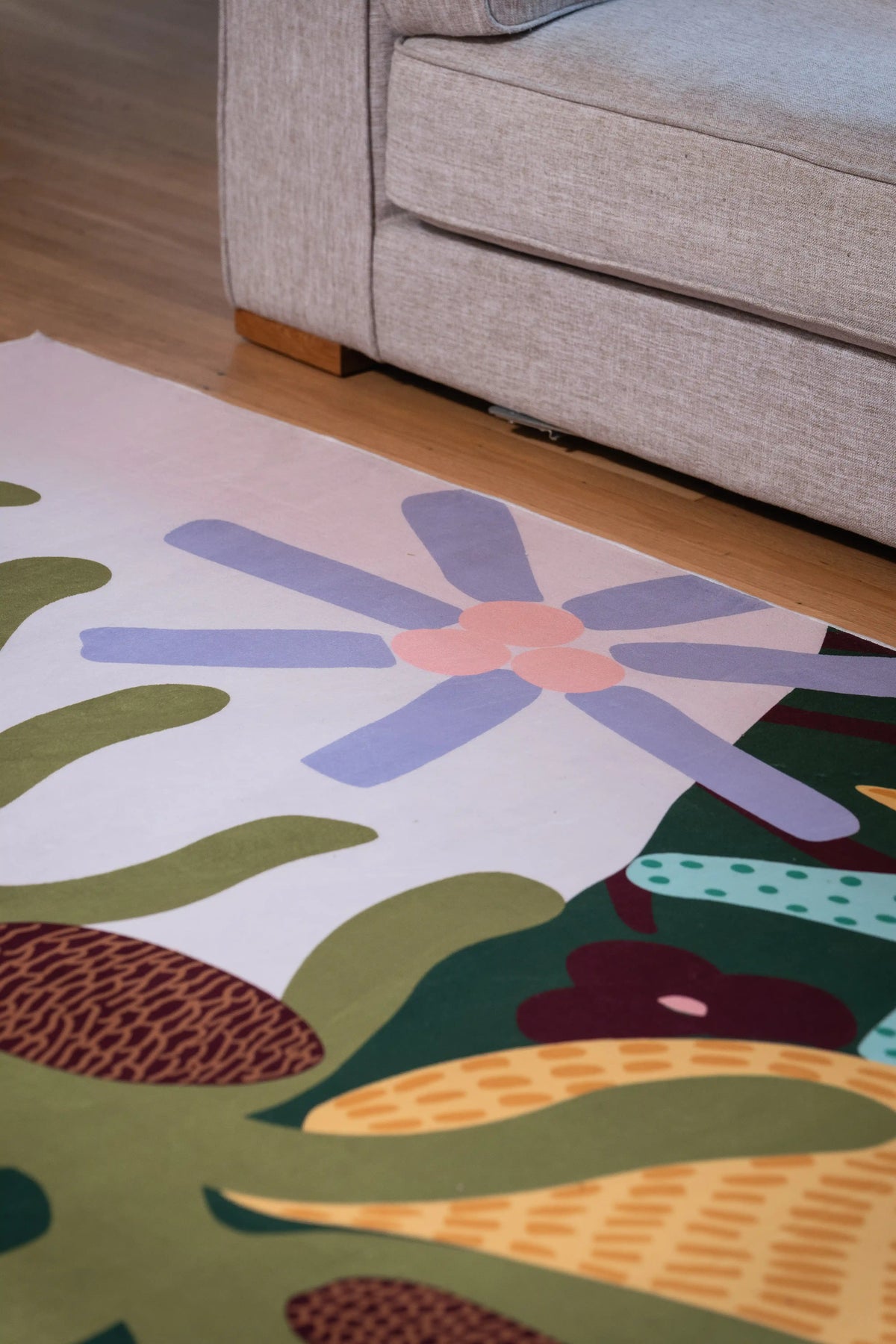 Lavender flower with a pink center, green leaves, and a brown textured pod along the rug edge beside a gray sofa.