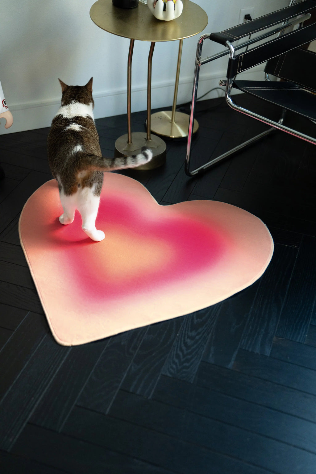Cat standing on the pink gradient heart rug beneath a round brass side table in a modern reading corner