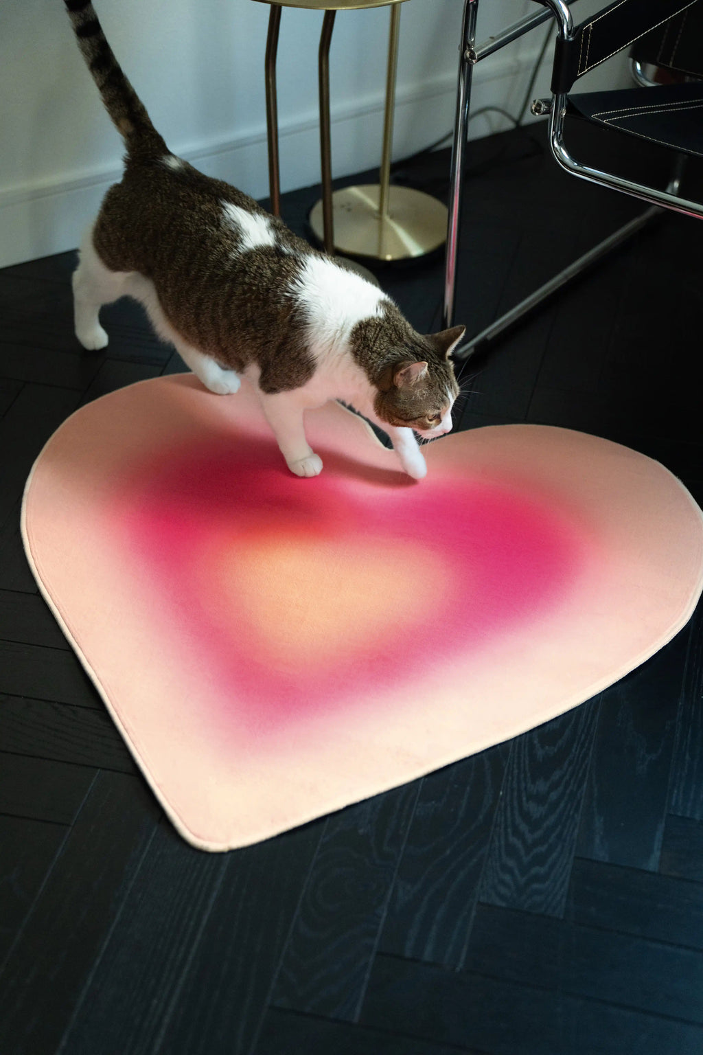 Cat walking across the Pink Modern Gradient Heart Rug beside a chrome chair on a dark herringbone floor