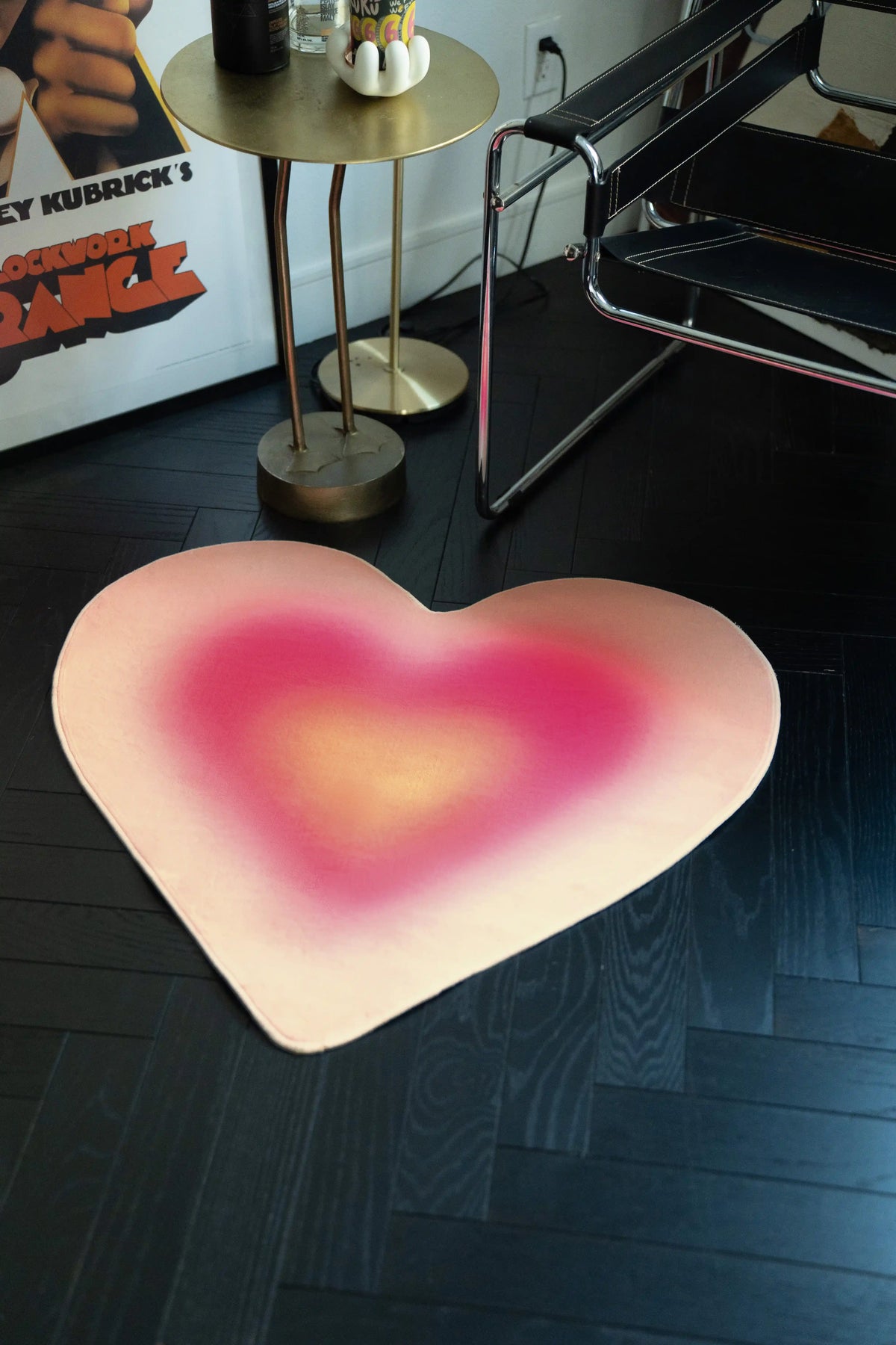 Heart-shaped pink rug on a dark wooden floor with a poster and small table in the background.