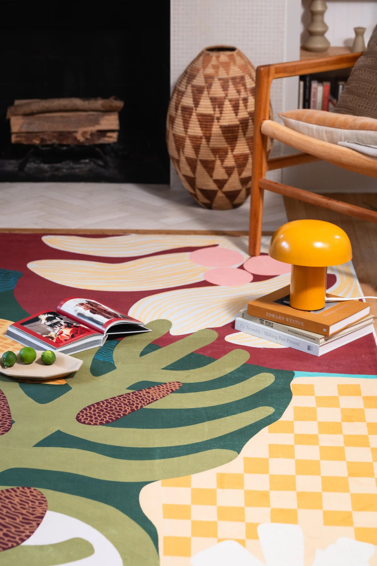 Cream flower, green leaves, and yellow checks in front of a fireplace beside a wooden chair, books, and yellow lamp.