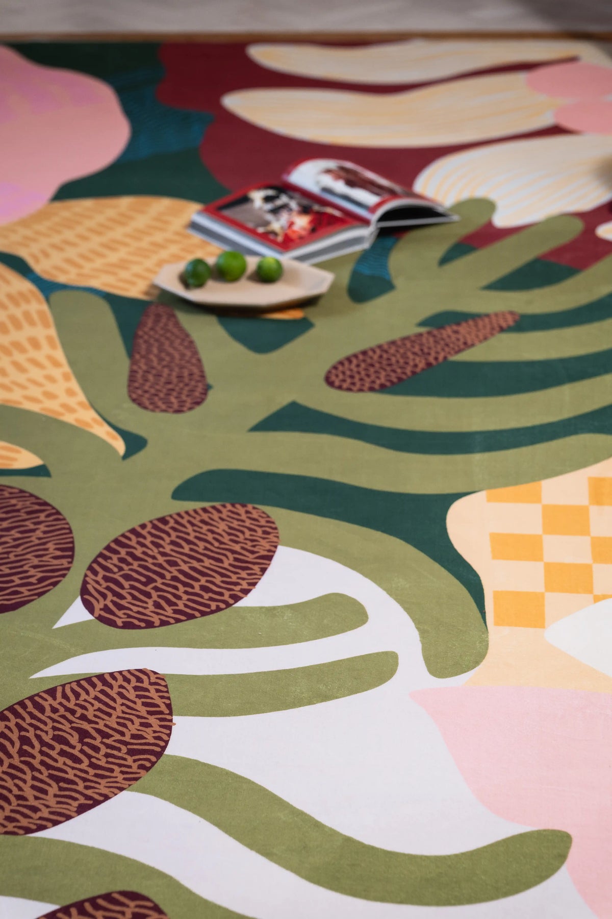 Green tropical leaves and brown seed pods across a pink and cream rug with a plate of green fruit near the top.