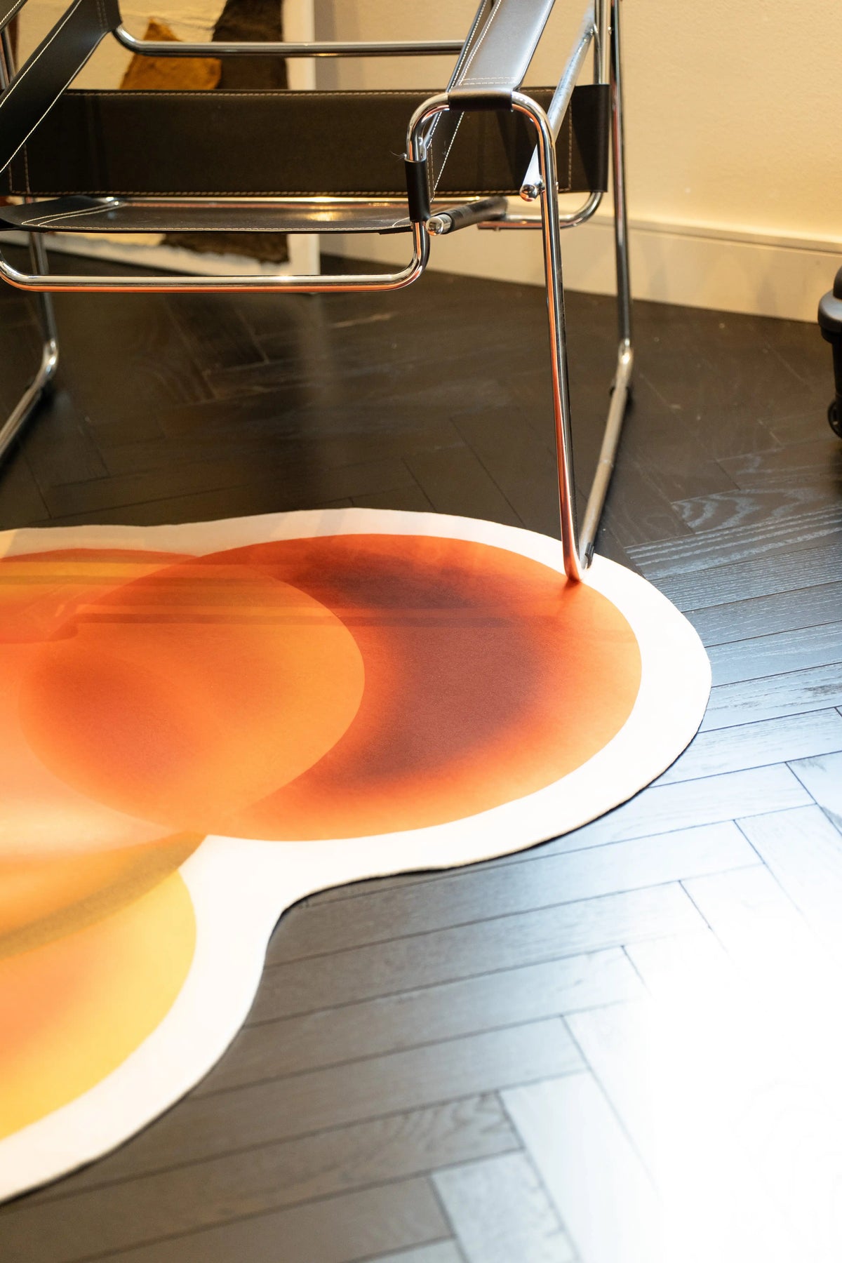 Earth tone rug beneath a chrome chair leg on a dark herringbone floor with warm circular color gradients