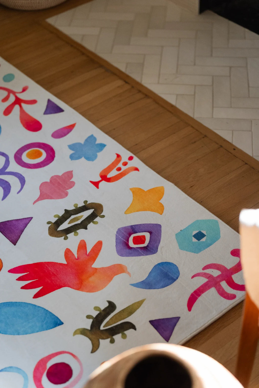 Corner close up of the graphic pattern rug near a cup on the table, showing bright nature inspired symbols on a white ground