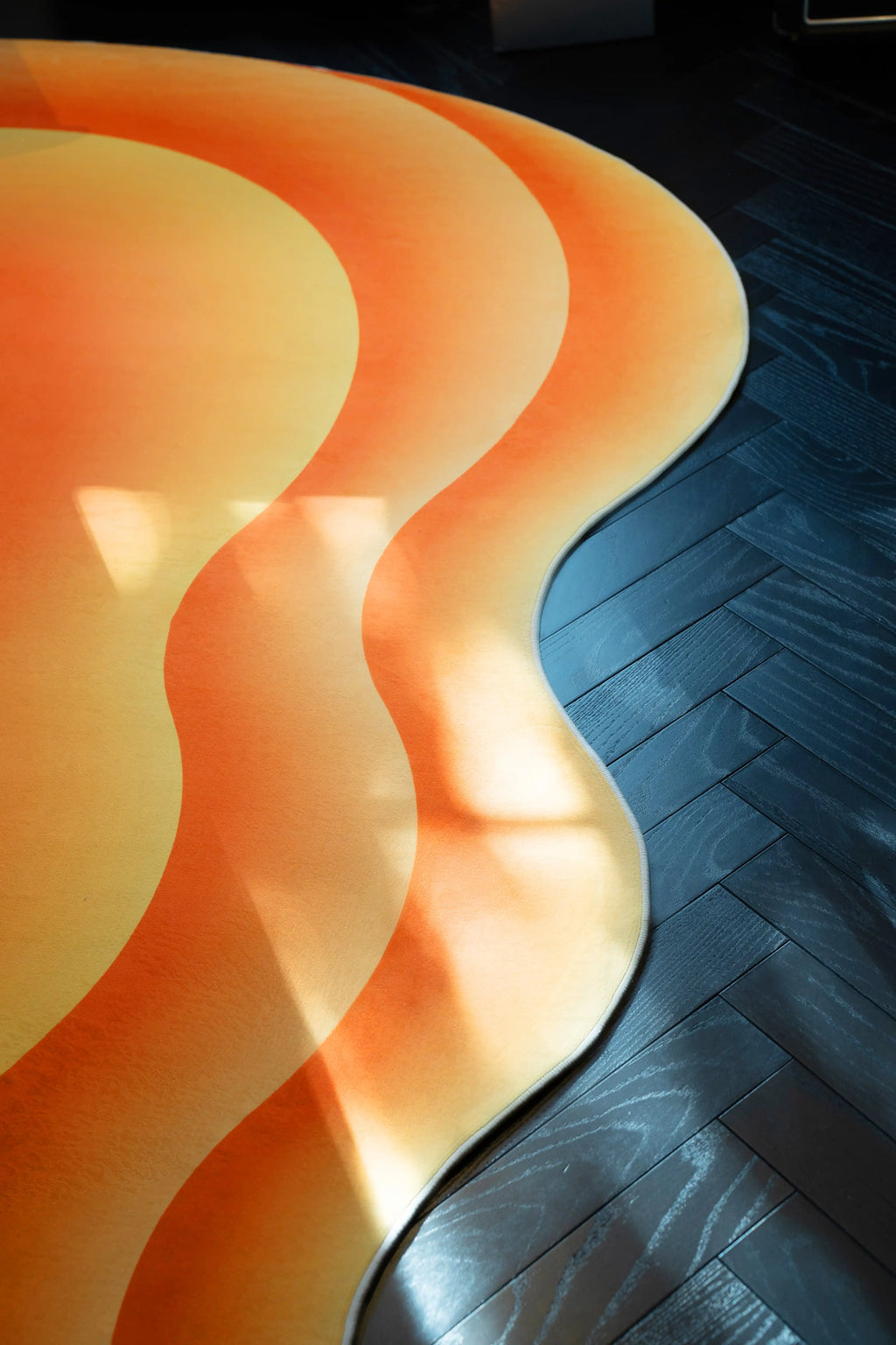 Side view of the wavy area rug with orange and yellow contours on a dark herringbone wood floor