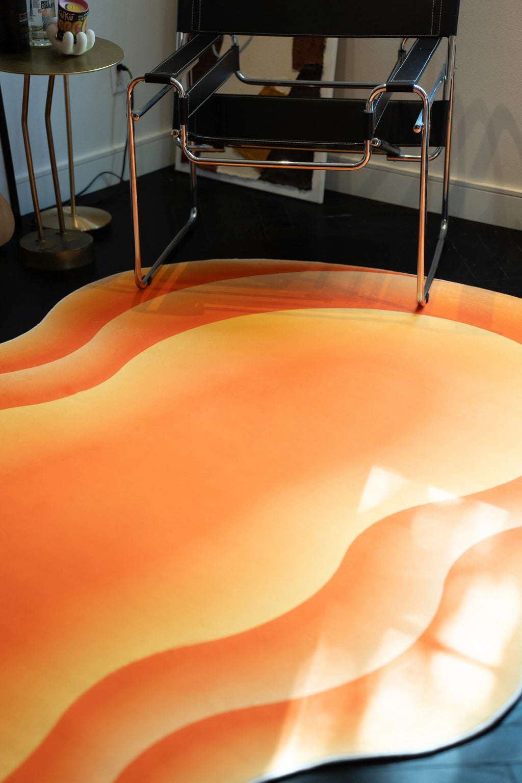 Close up of the orange abstract rug beneath a chrome chair, showing layered wavy bands and soft sunlight