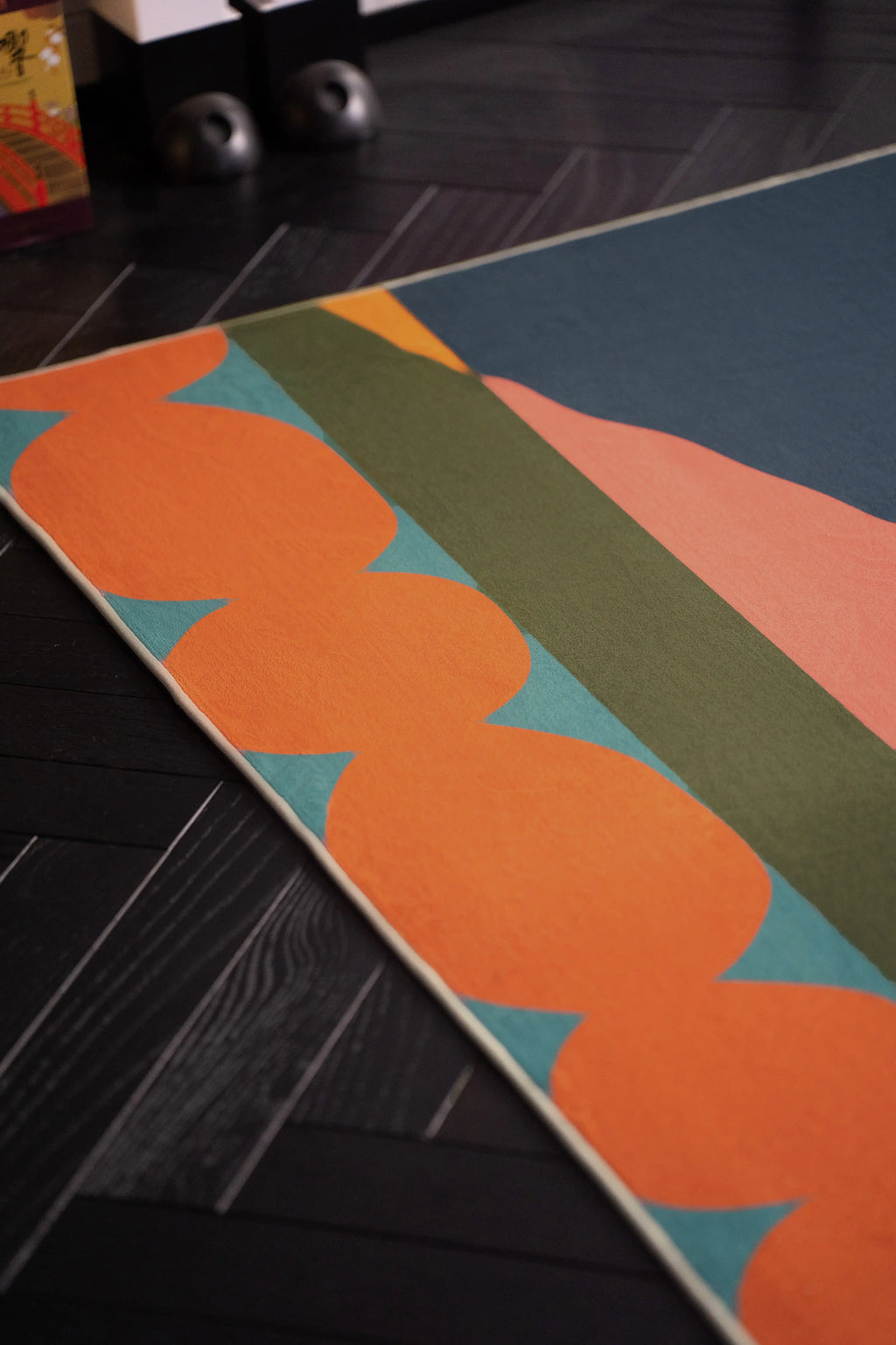 Partial view of the landscape rug with layered blue, olive, coral, and cream shapes on a dark herringbone floor