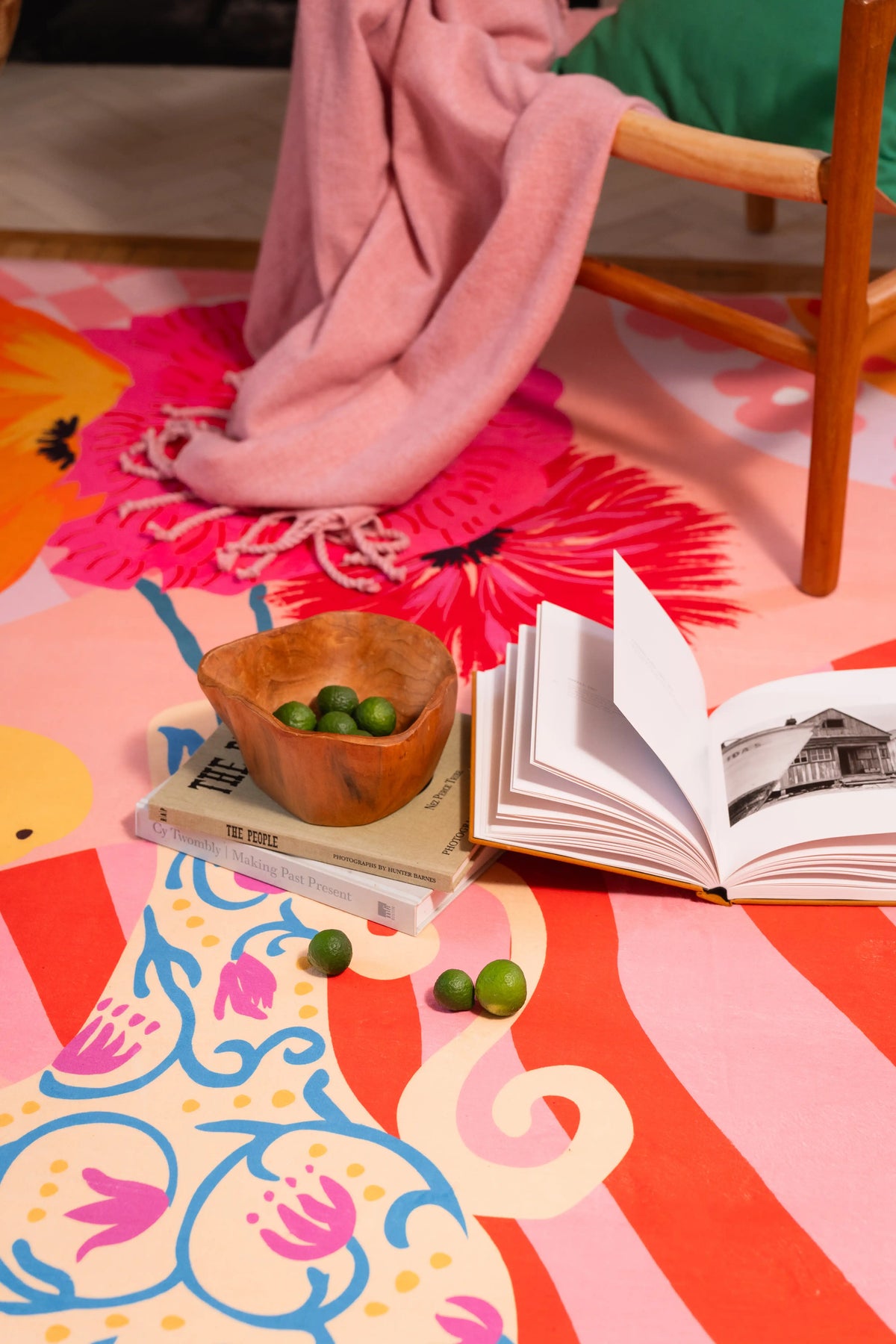Floral and striped rug beneath a wooden chair with a pink throw, open book, wooden bowl, and green fruit.