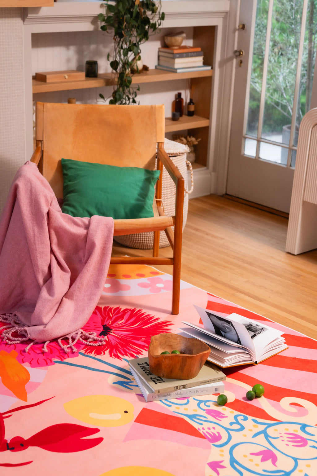Colorful rug beside a wooden chair with a green cushion and pink throw, with books and a wooden bowl on top.