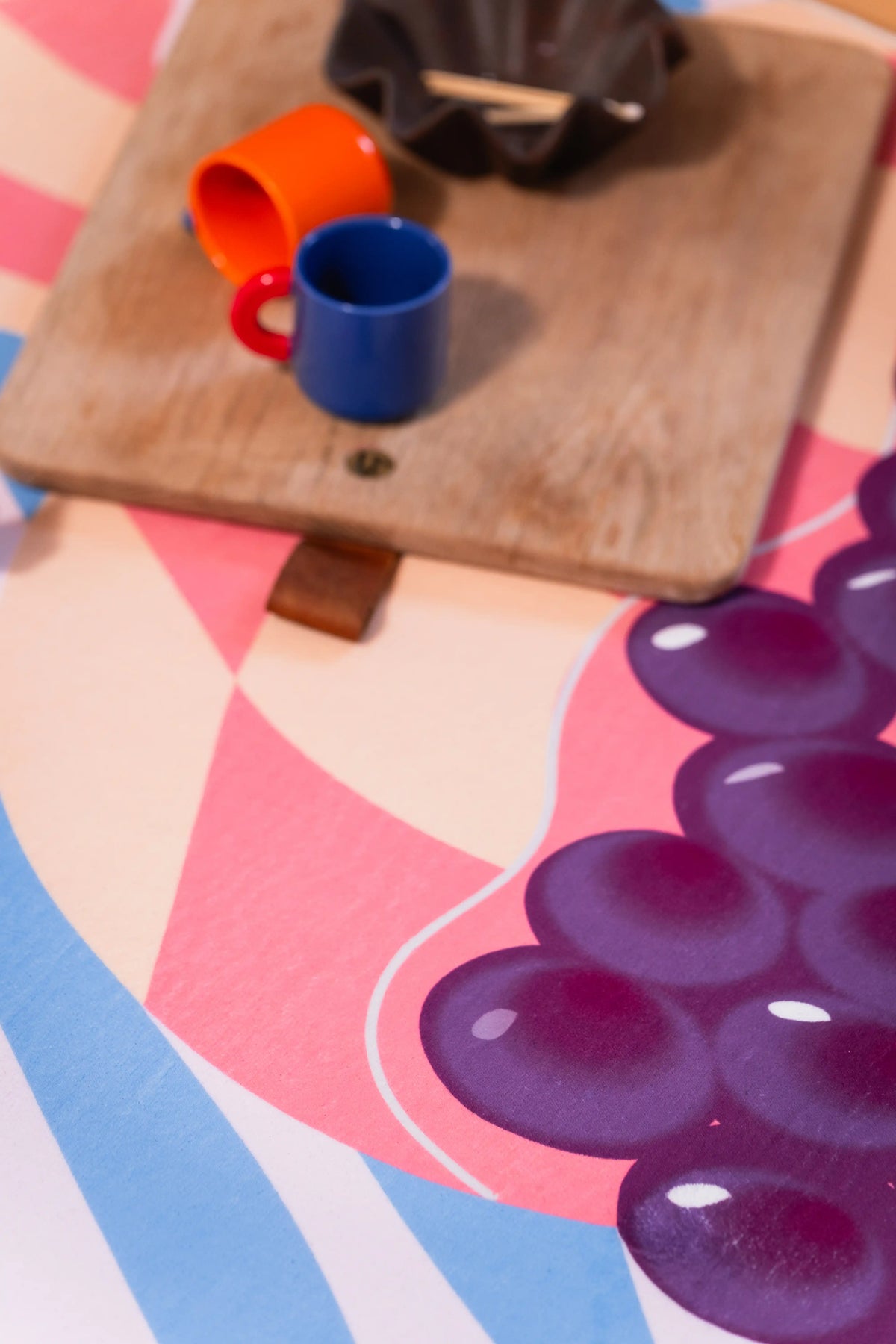 Orange and blue cups on a wooden board beside purple grapes and pink geometric shapes on the rug.