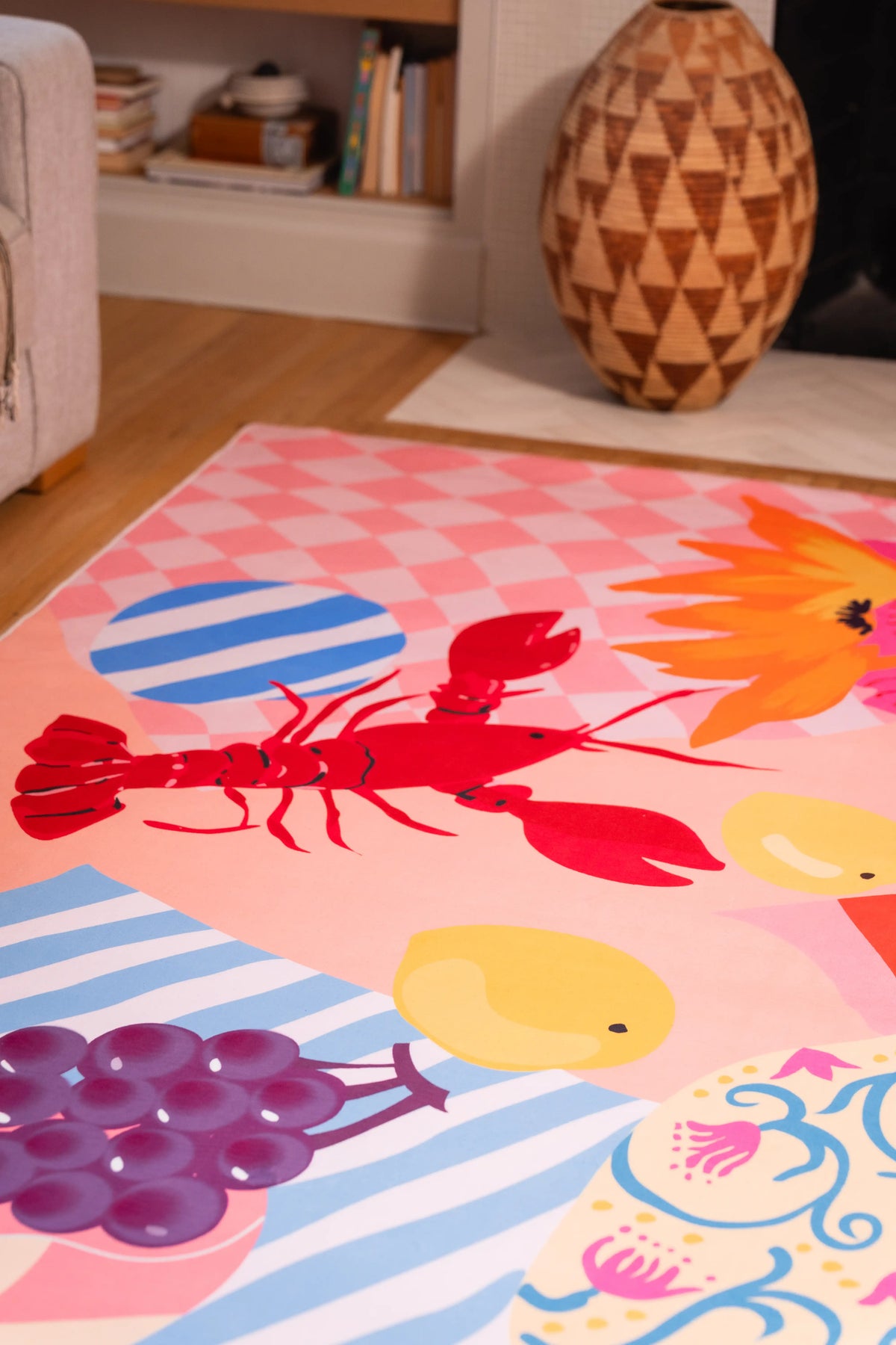 Red lobster, purple grapes, yellow lemons, blue-striped plate, and floral motifs on a pink rug near a carved wooden vase.