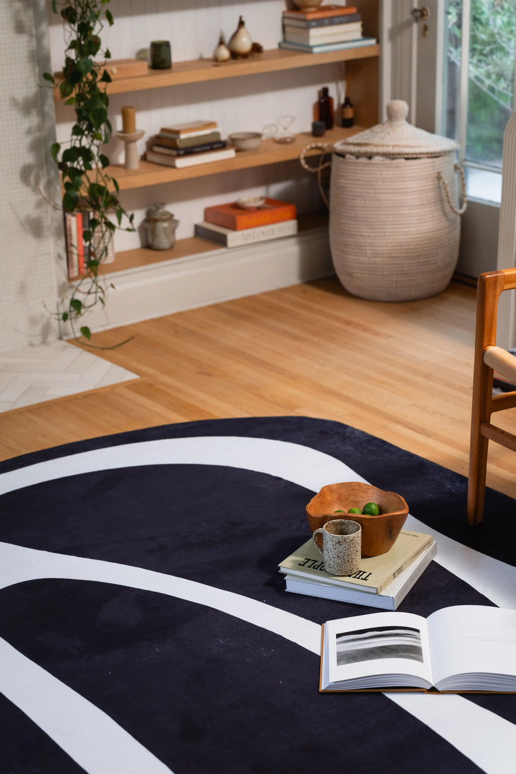 Wide white curve across a black rug beside a wooden chair, open book, and bowl on stacked books.