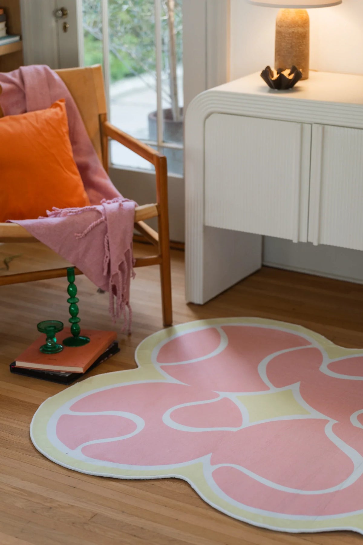 Pink tennis rug beside a white cabinet and wood chair with an orange pillow in a casual corner