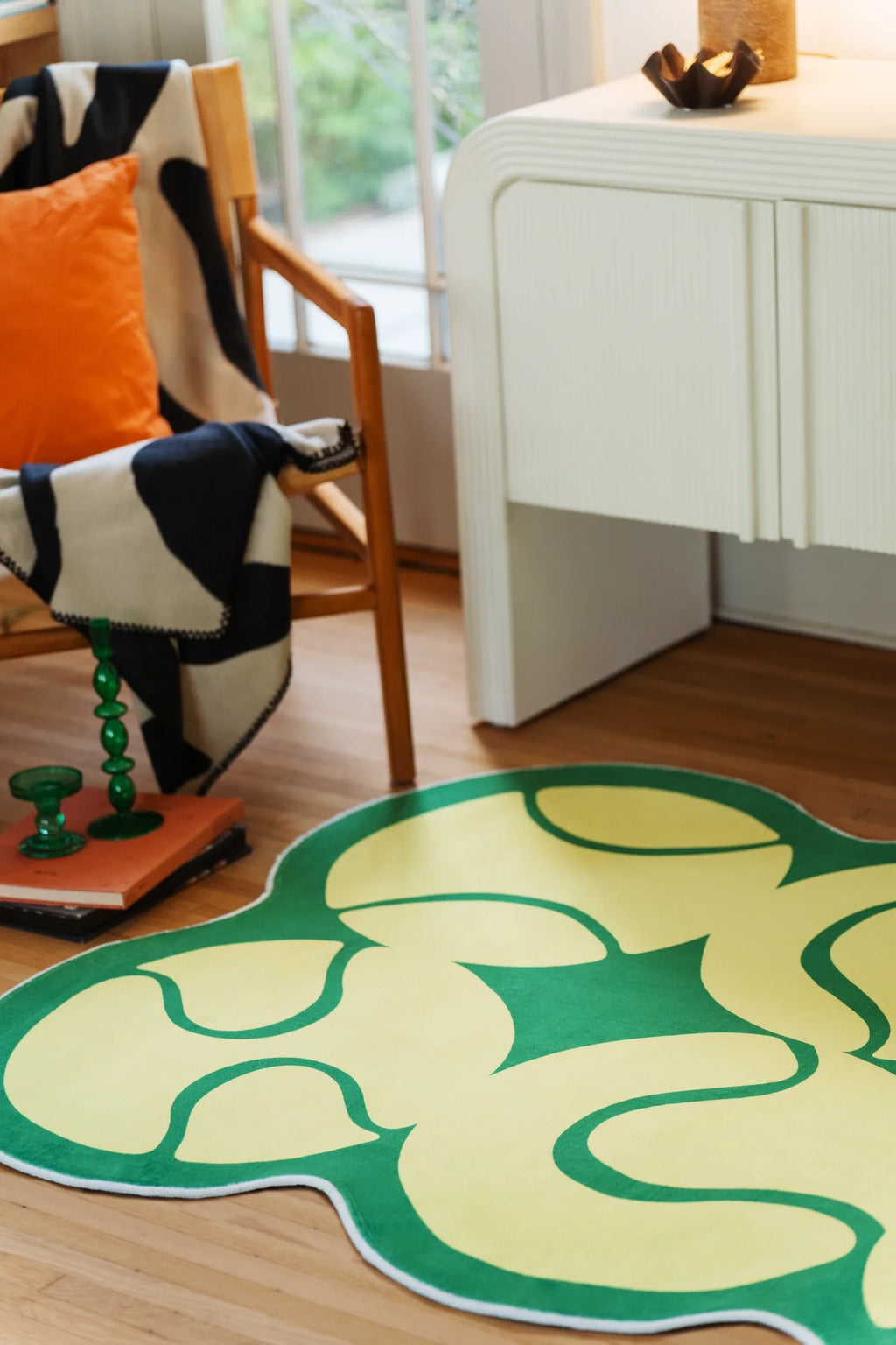 Yellow irregular rug beside a white media console and wood chair, with curved green lines across the surface