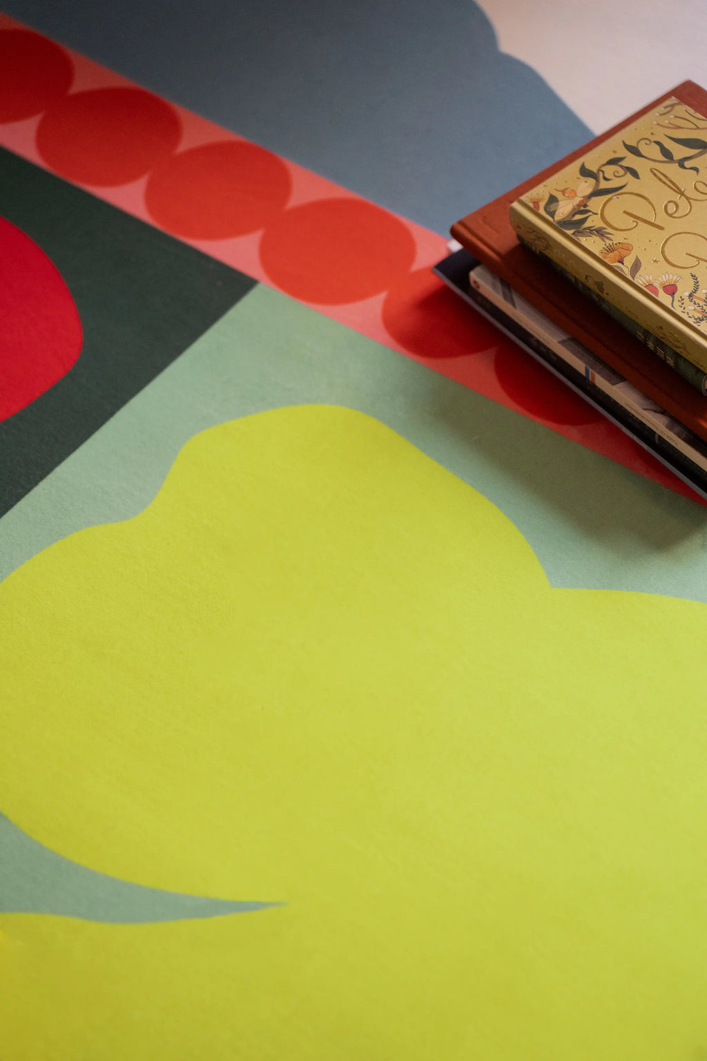 Close up of the flower rug with a lime green shape, red petal form, orange dots, and stacked books at the corner
