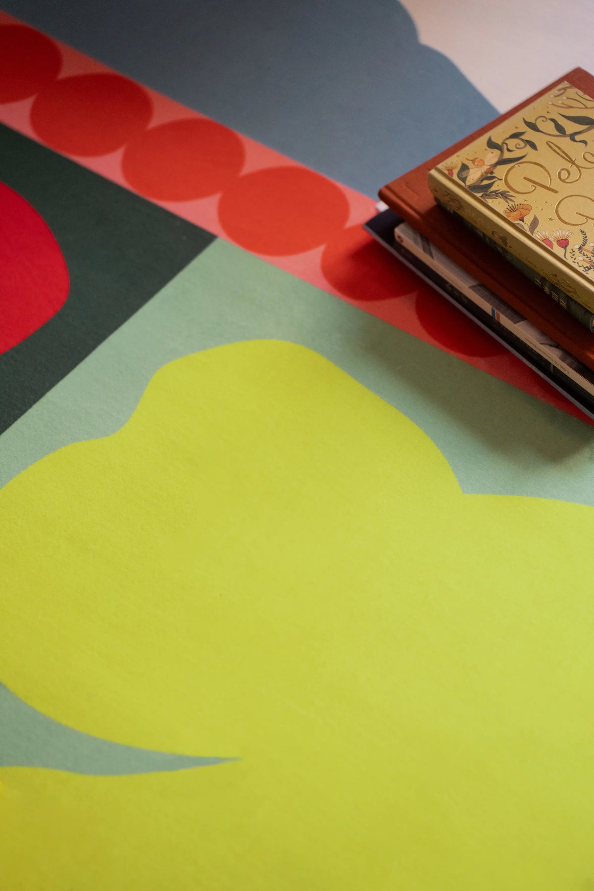 Close up of the flower rug with a lime green shape, red petal form, orange dots, and stacked books at the corner
