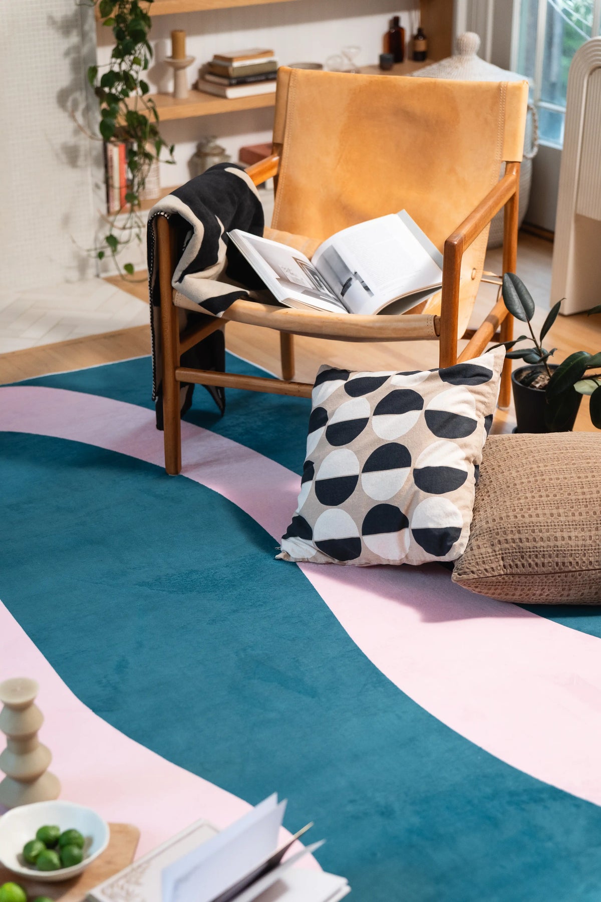 A wooden lounge chair and black-and-white floor cushion overlap the edge of the blue and pink rug.