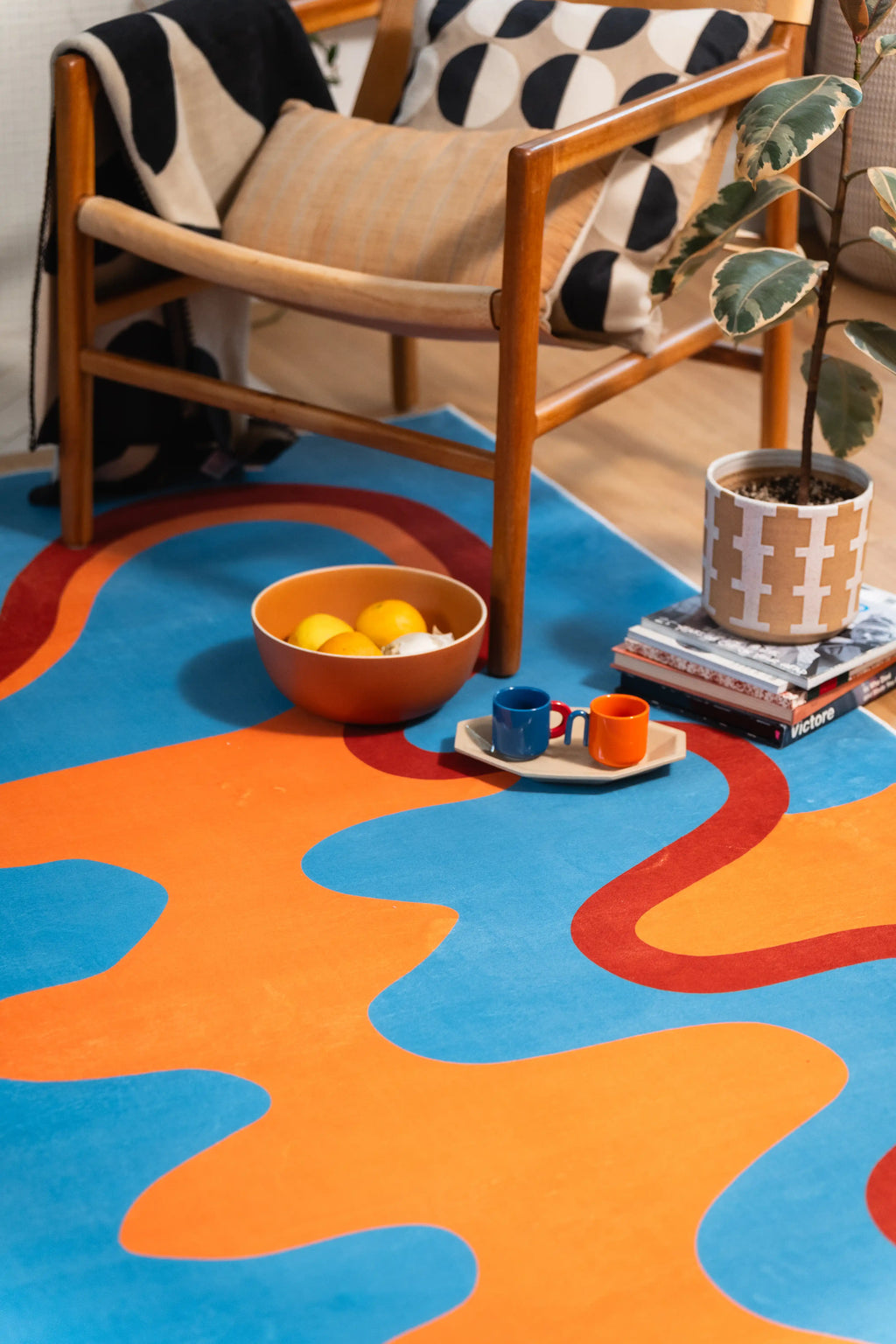 A styled corner of the wave rug beneath a wooden chair, with an orange bowl, cups, and stacked books.