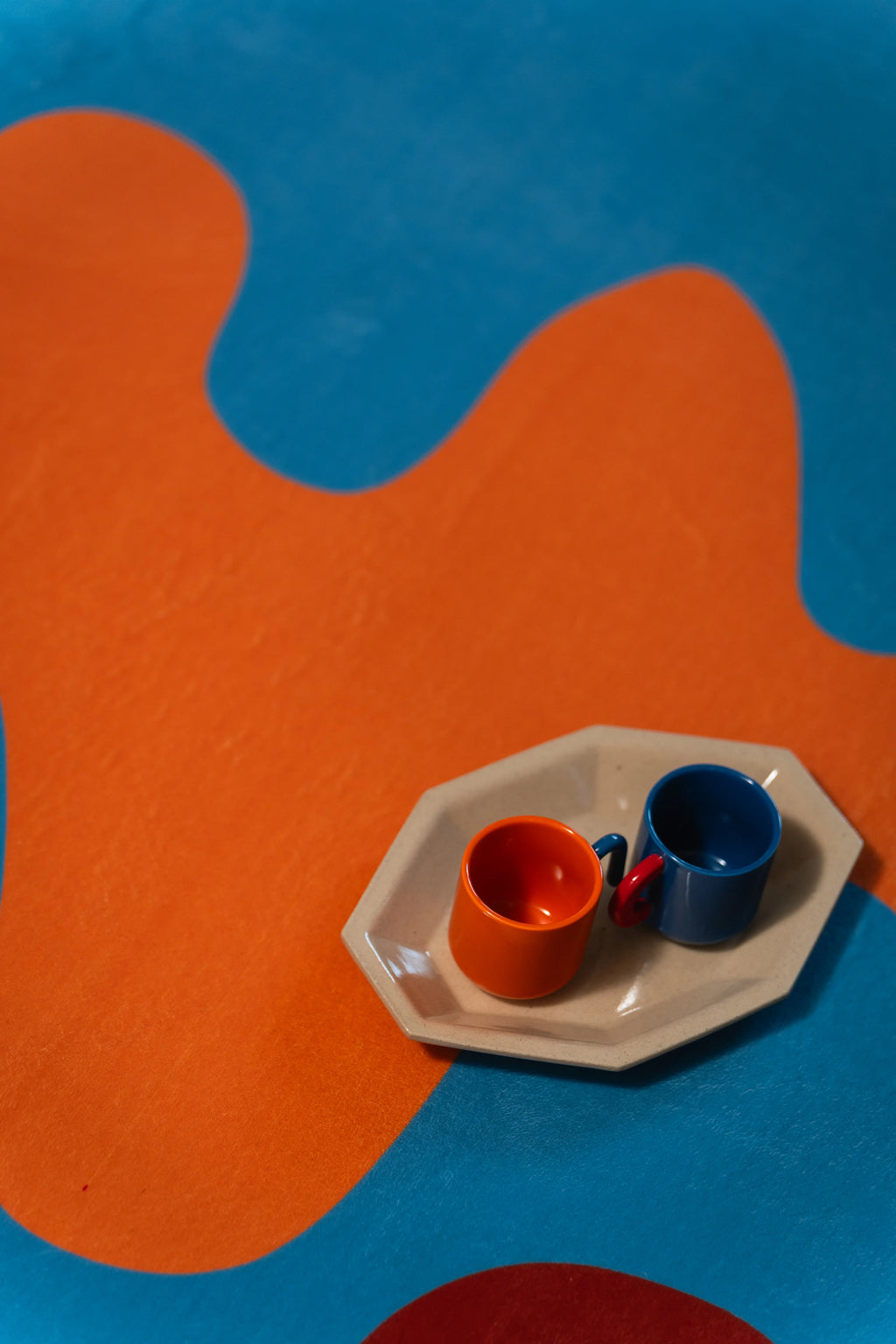 Two colored cups on a small tray rest on an orange wave surrounded by blue and a curved red line.