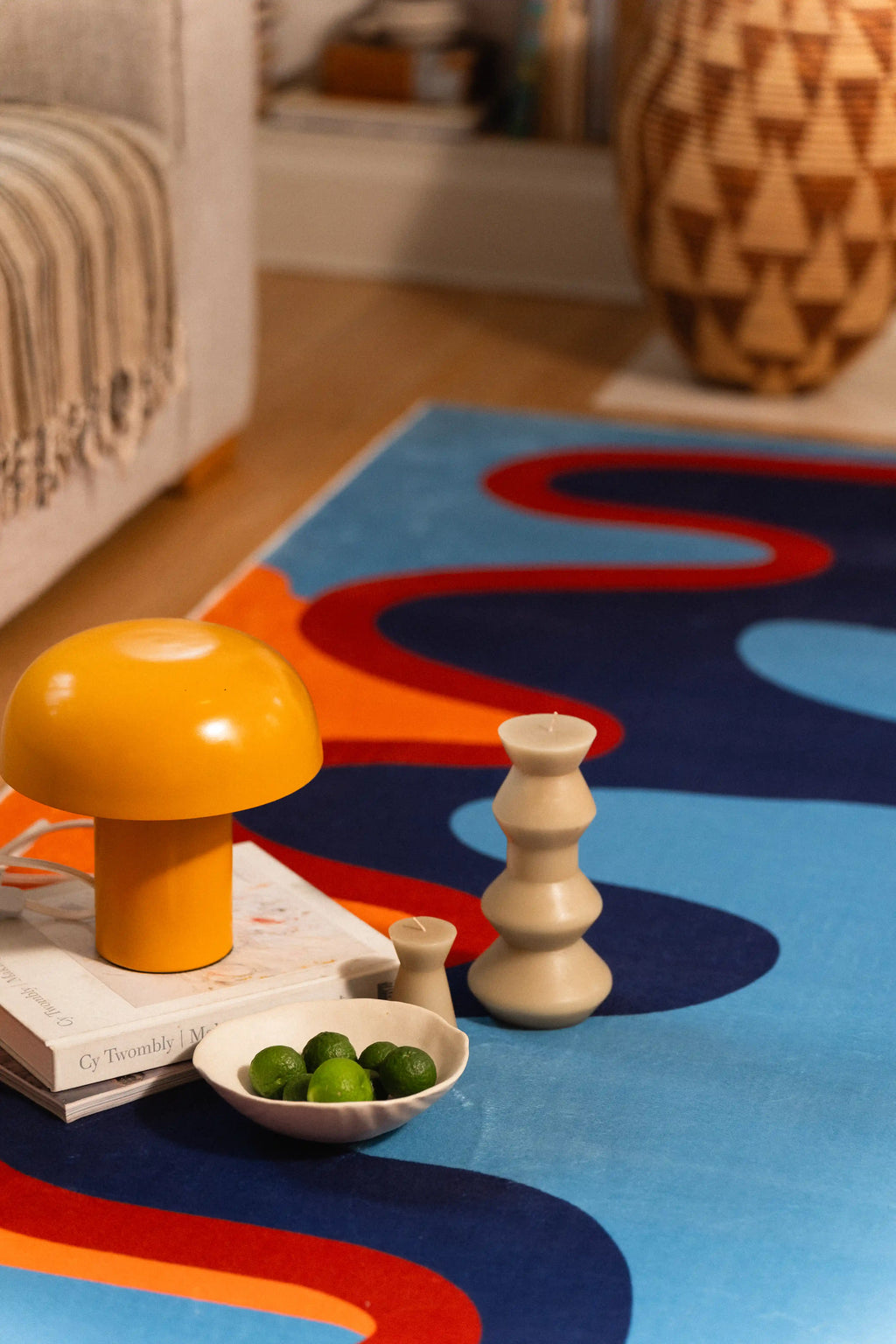 An orange lamp, white sculptural candle, and bowl of green fruit rest beside navy shapes on the blue rug.