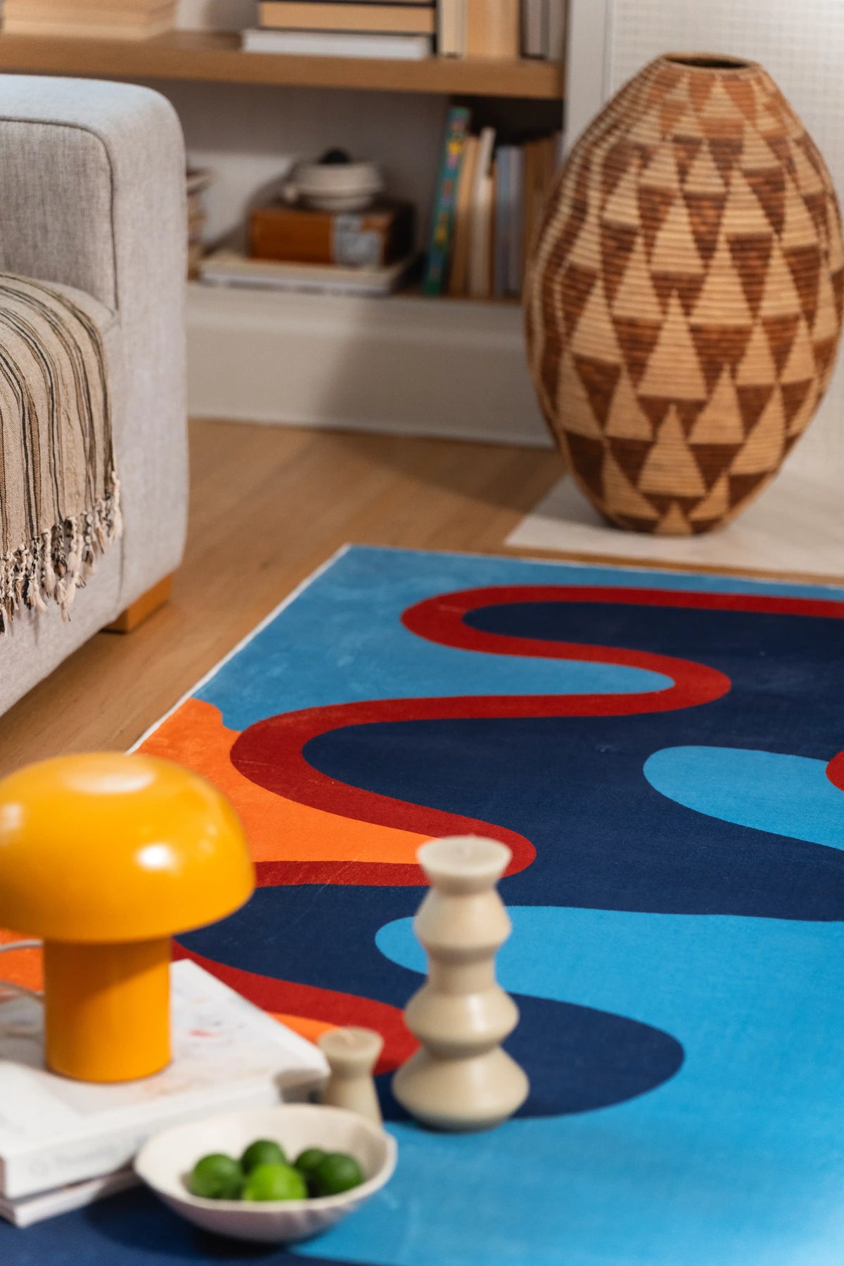 A rust-red loop crosses the blue field behind an orange lamp, white candle, and bowl of green fruit.