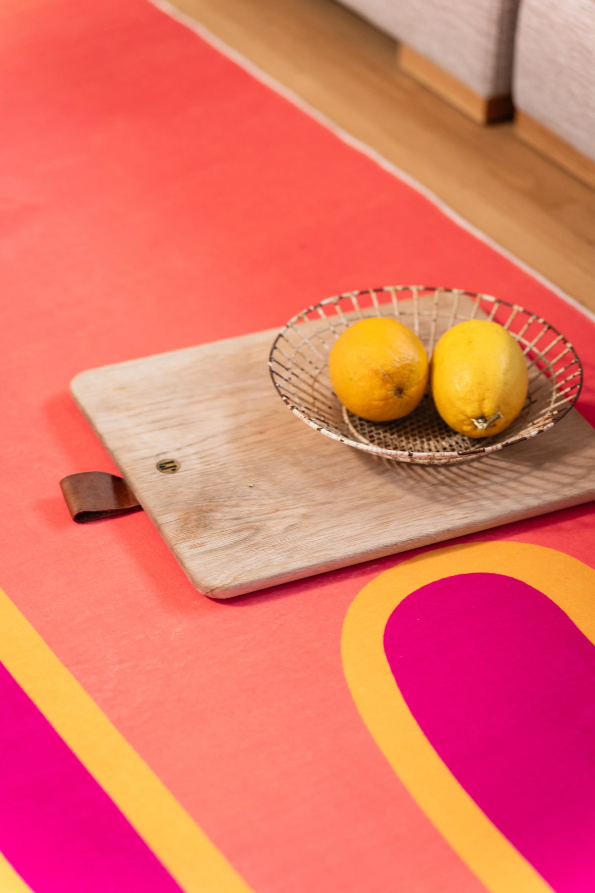 Wooden board and wire bowl filled with lemons resting on a coral rug beside its magenta stripe.