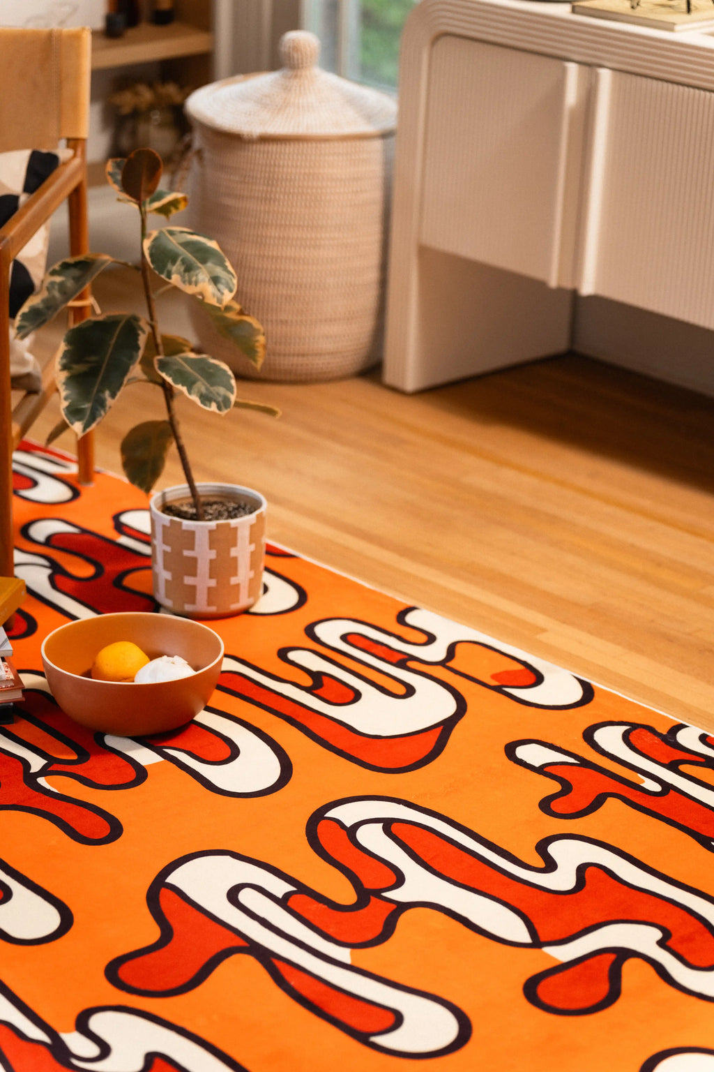 Orange abstract rug beside a potted plant and white cabinet on a light wood floor.