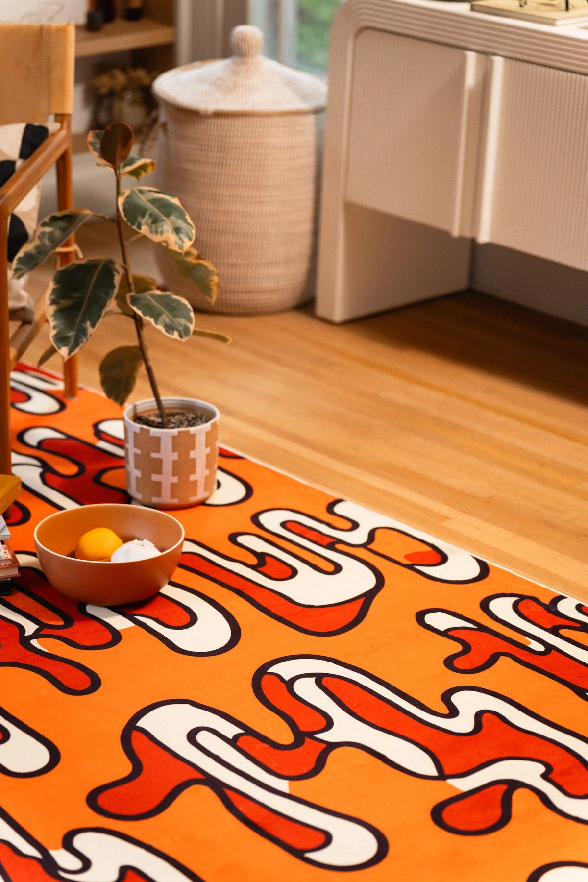 Orange abstract rug beside a potted plant and white cabinet on a light wood floor.