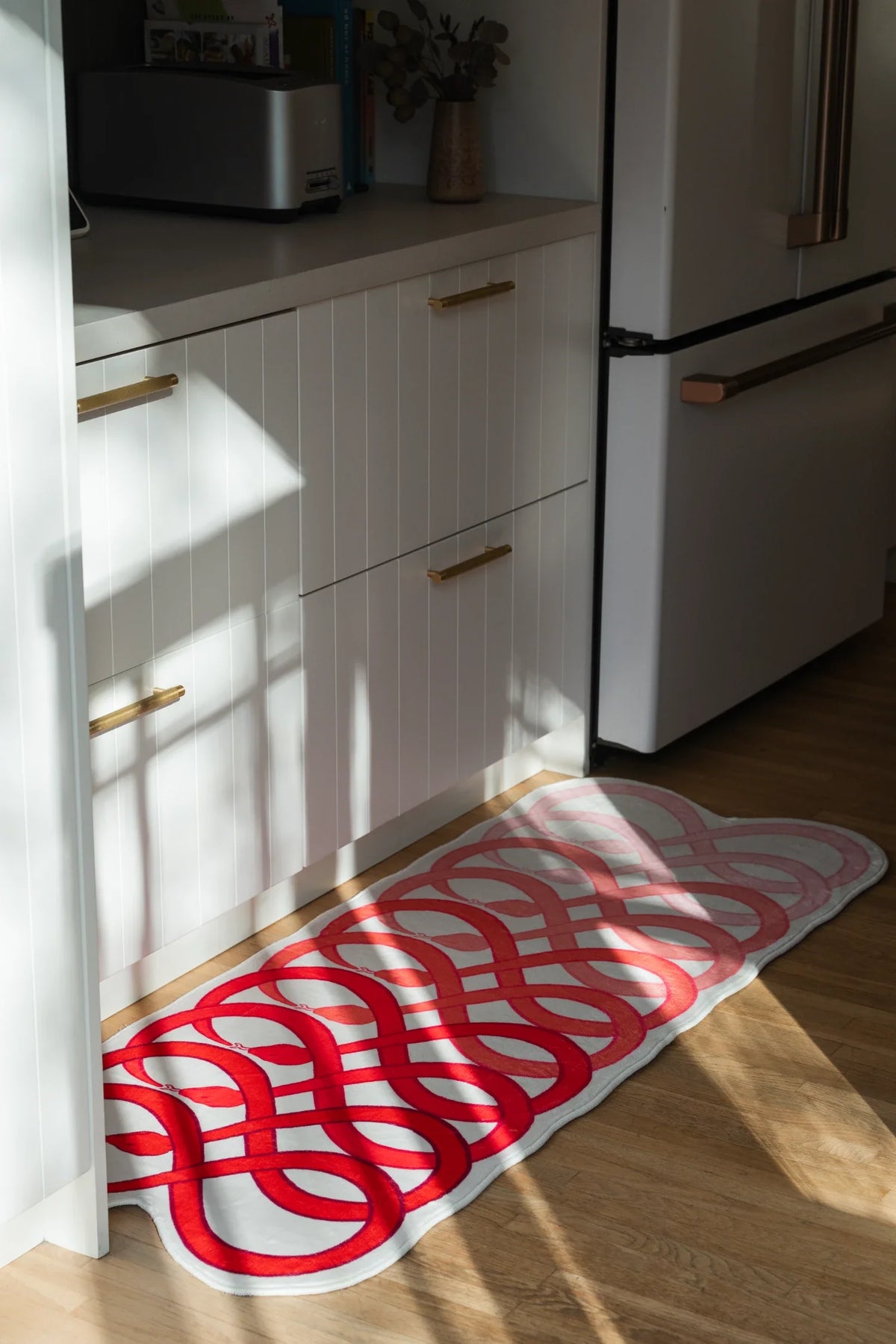 Red runner rug laid along a narrow kitchen path beside white cabinets and a refrigerator on a light wood floor