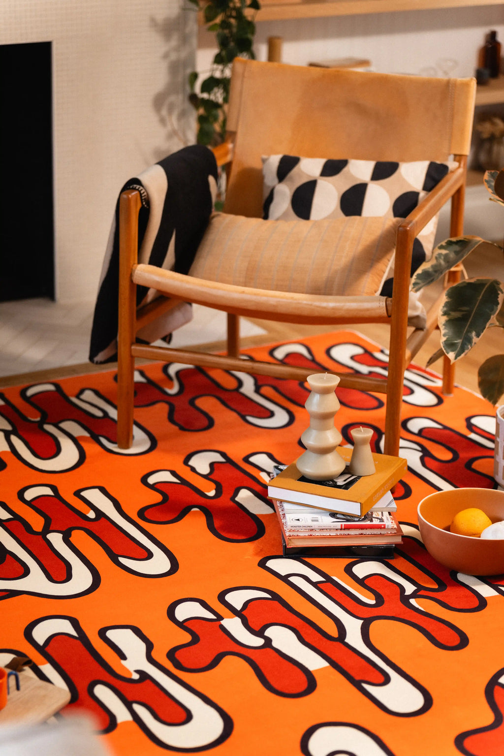 Orange graphic rug beneath a wooden armchair with books, a candle, and a bowl arranged in front.
