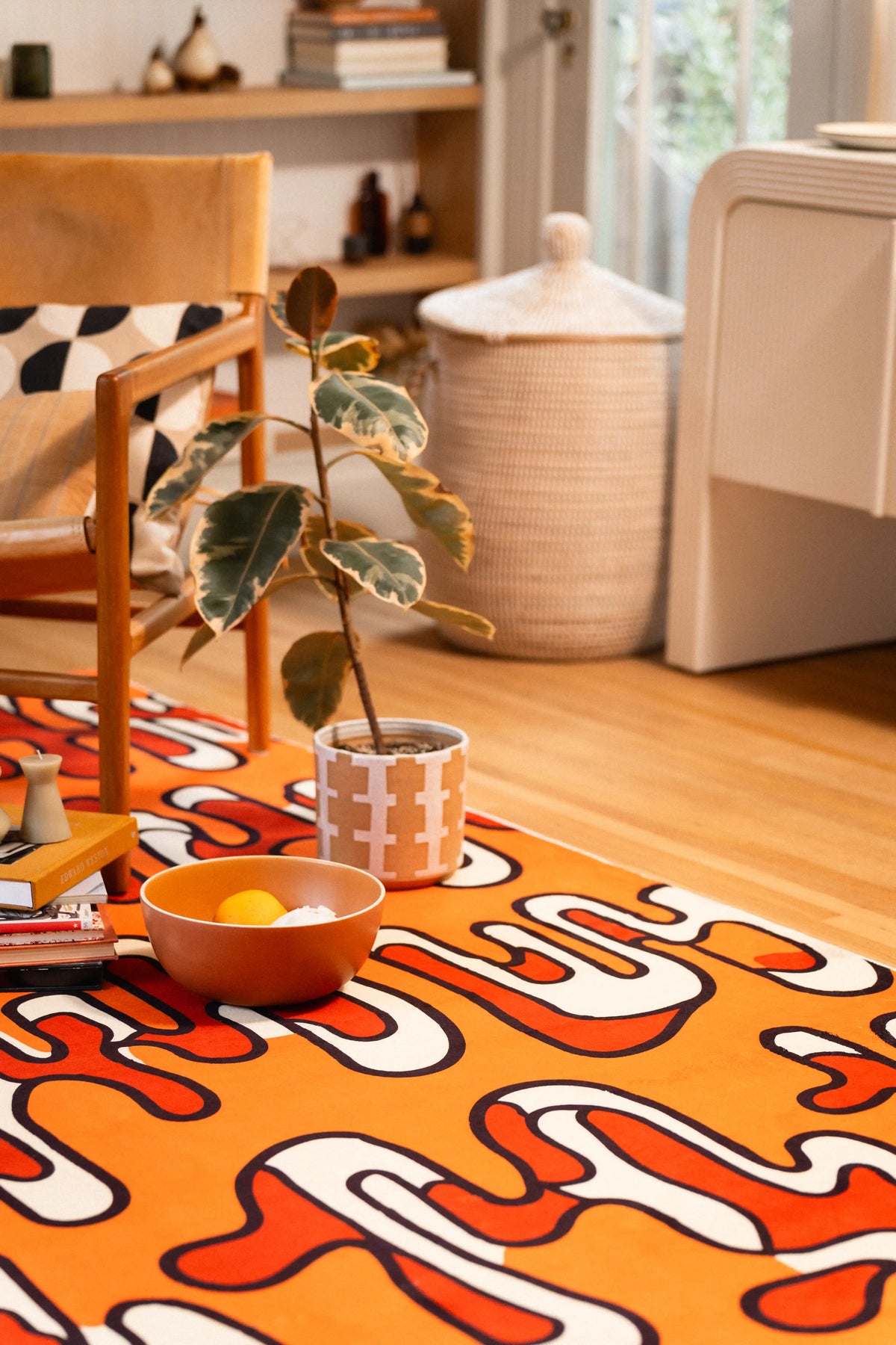 Orange abstract area rug with a potted plant, bowl, and patterned mug beside a wooden armchair.