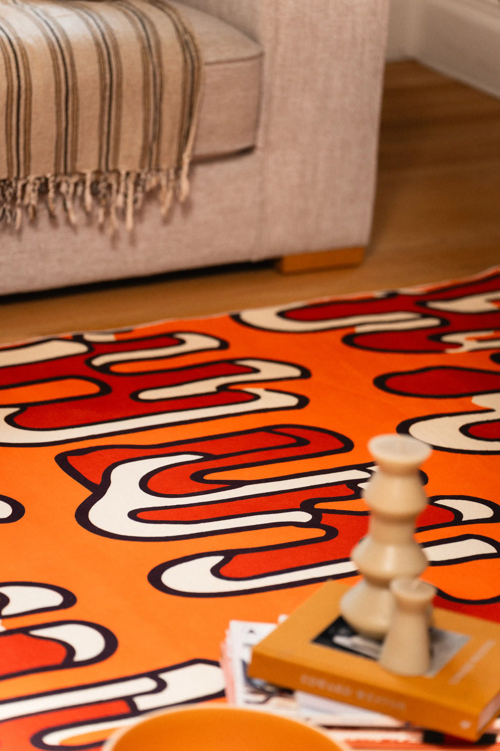 Close-up of an orange squiggle rug beside a gray sofa with stacked books and a sculptural candle.