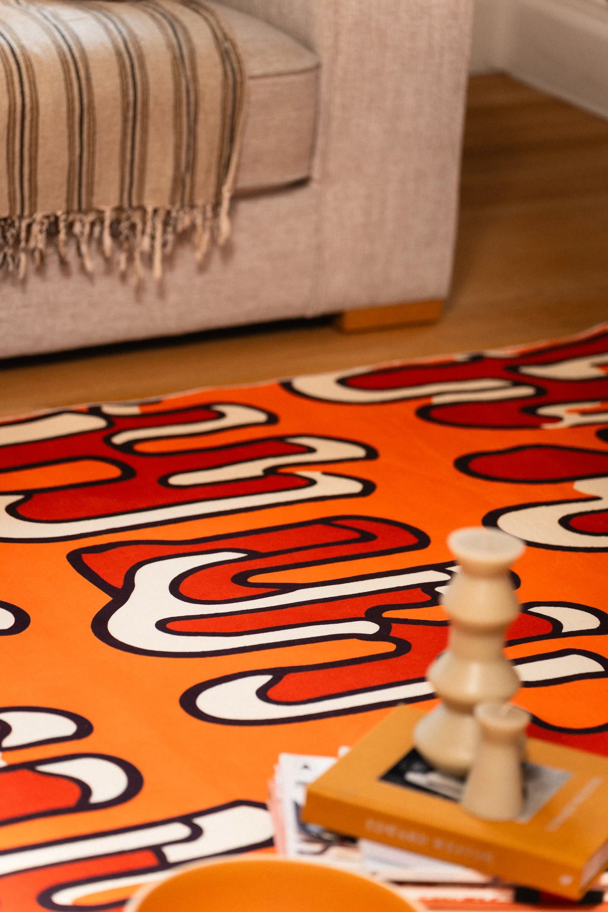 Close-up of an orange squiggle rug beside a gray sofa with stacked books and a sculptural candle.