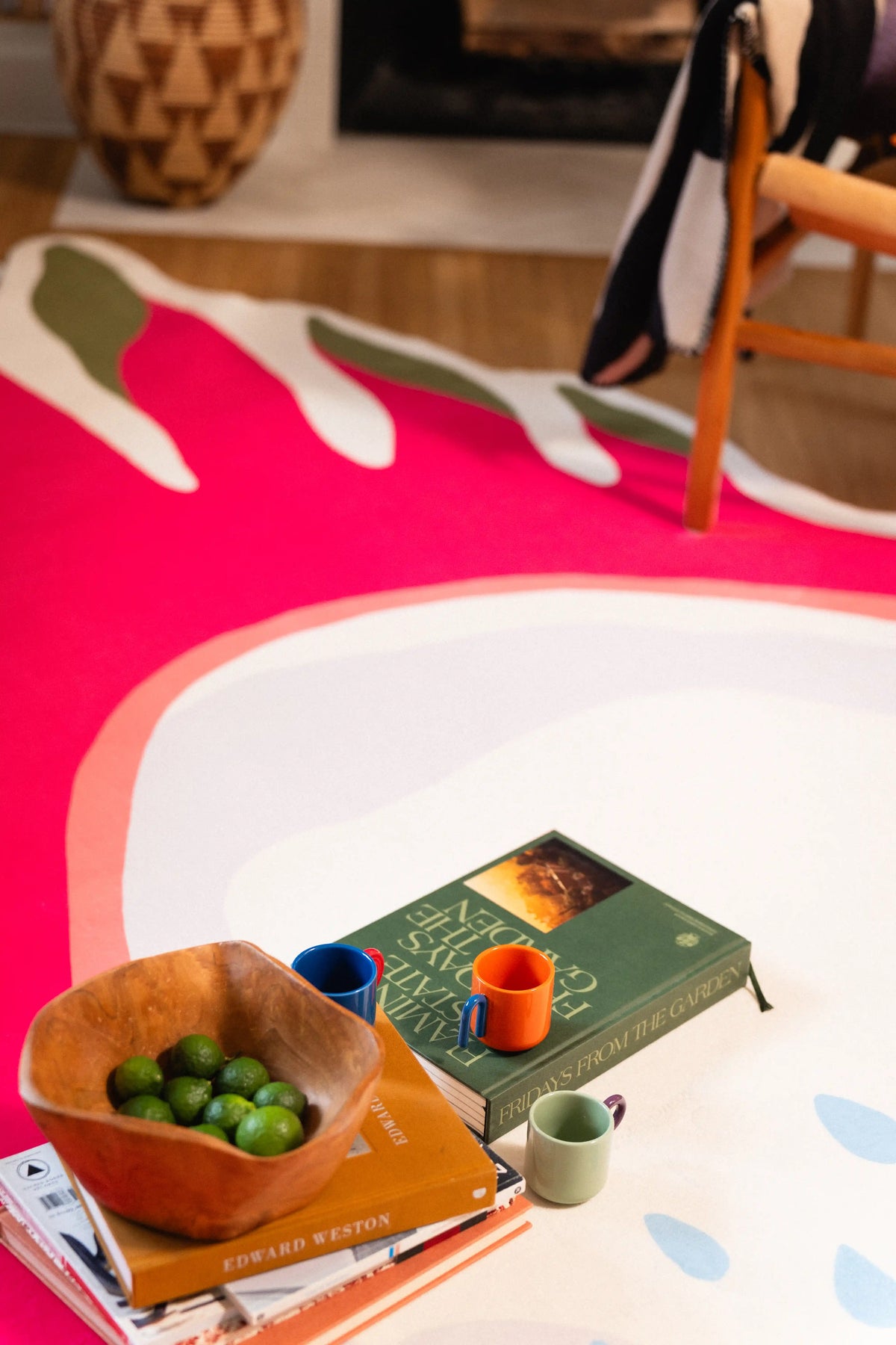 Wooden bowl of green fruit, stacked books, and blue, orange, and green cups on the pale center of a dragon fruit rug.