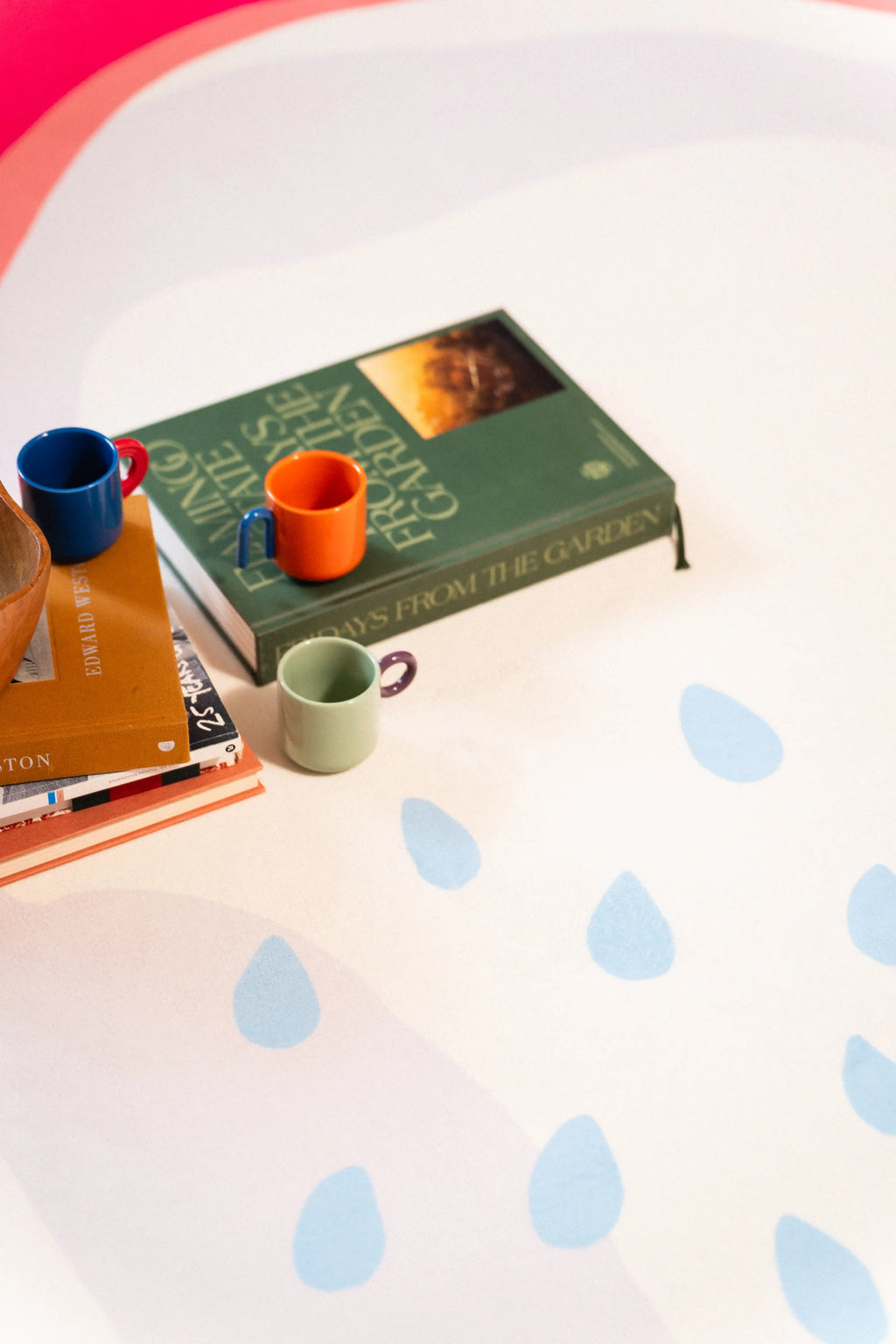Light blue seed shapes beside a green book, stacked books, wooden fruit bowl, and three small cups.
