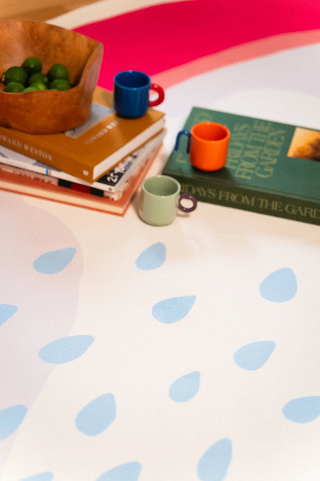 Light blue seed shapes beside stacked books, a green book, wooden fruit bowl, and colorful cups on the rug.