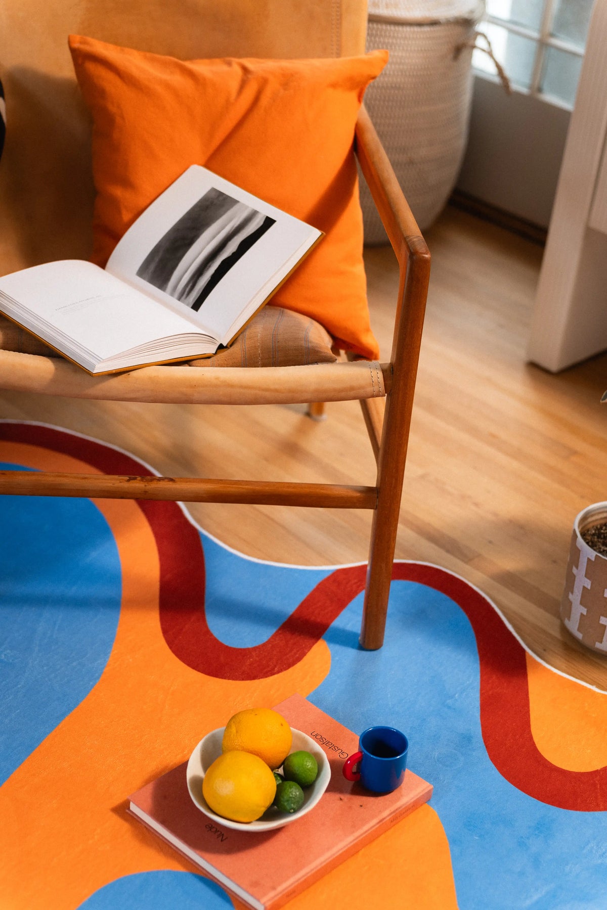 An open book rests on an orange-cushioned chair above rust-trimmed blue waves, lemons, and a small blue cup.