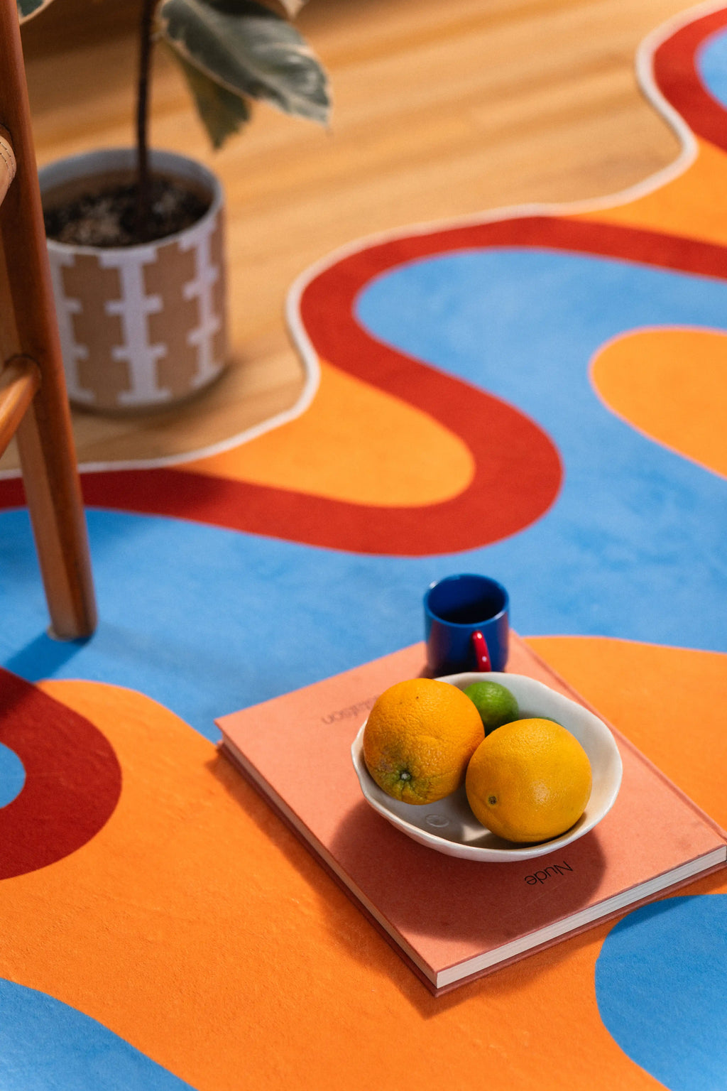 Sky-blue and orange shapes meet along a rust-red curve beside a potted plant and tray of lemons.