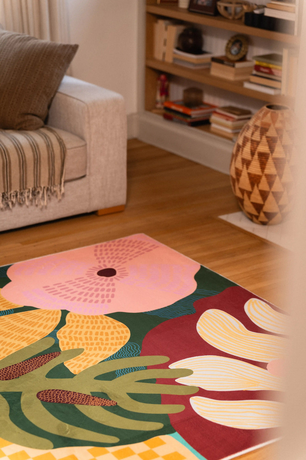 Pink and cream tropical flowers with green leaves on a rug beside a gray sofa and carved wooden vase.