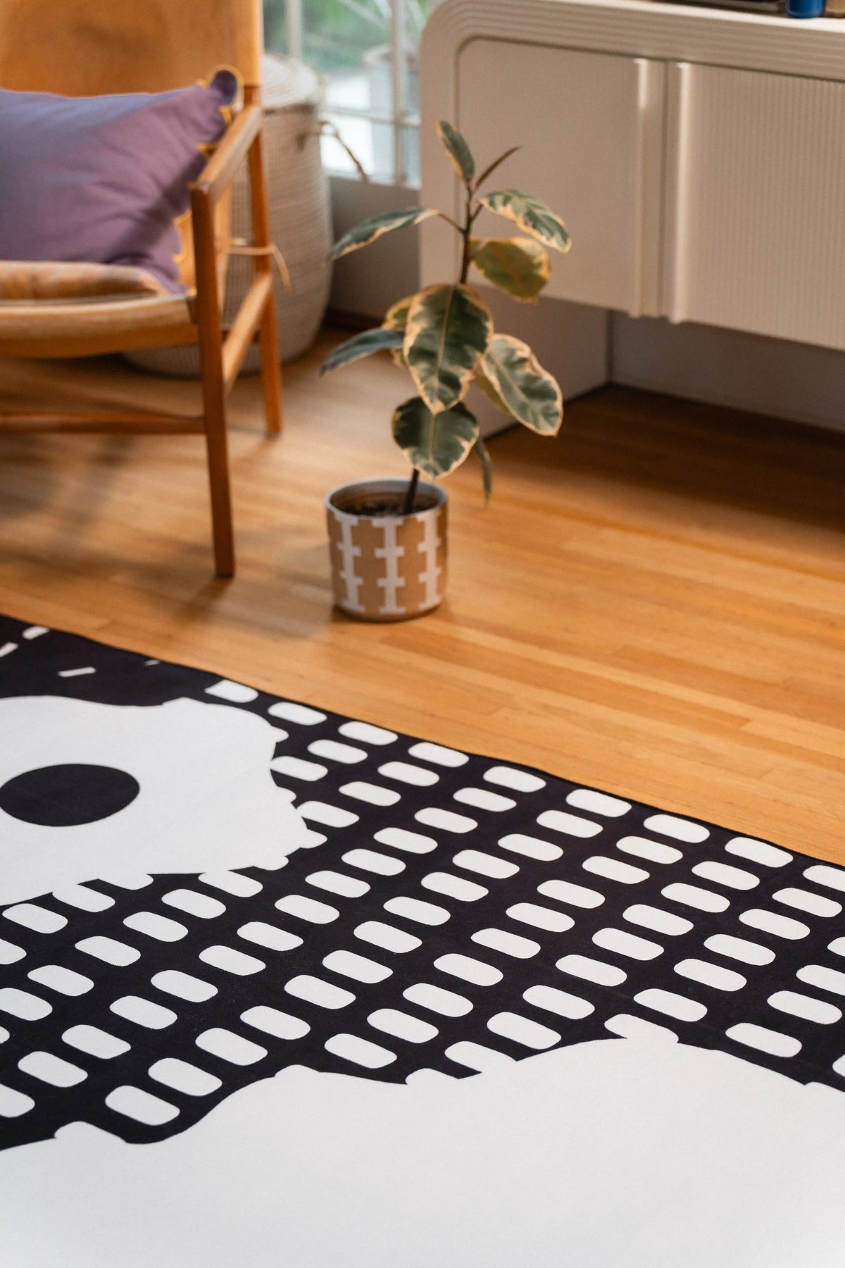 White flower shapes and rows of black grid marks on a rug beside a wooden chair and potted plant.
