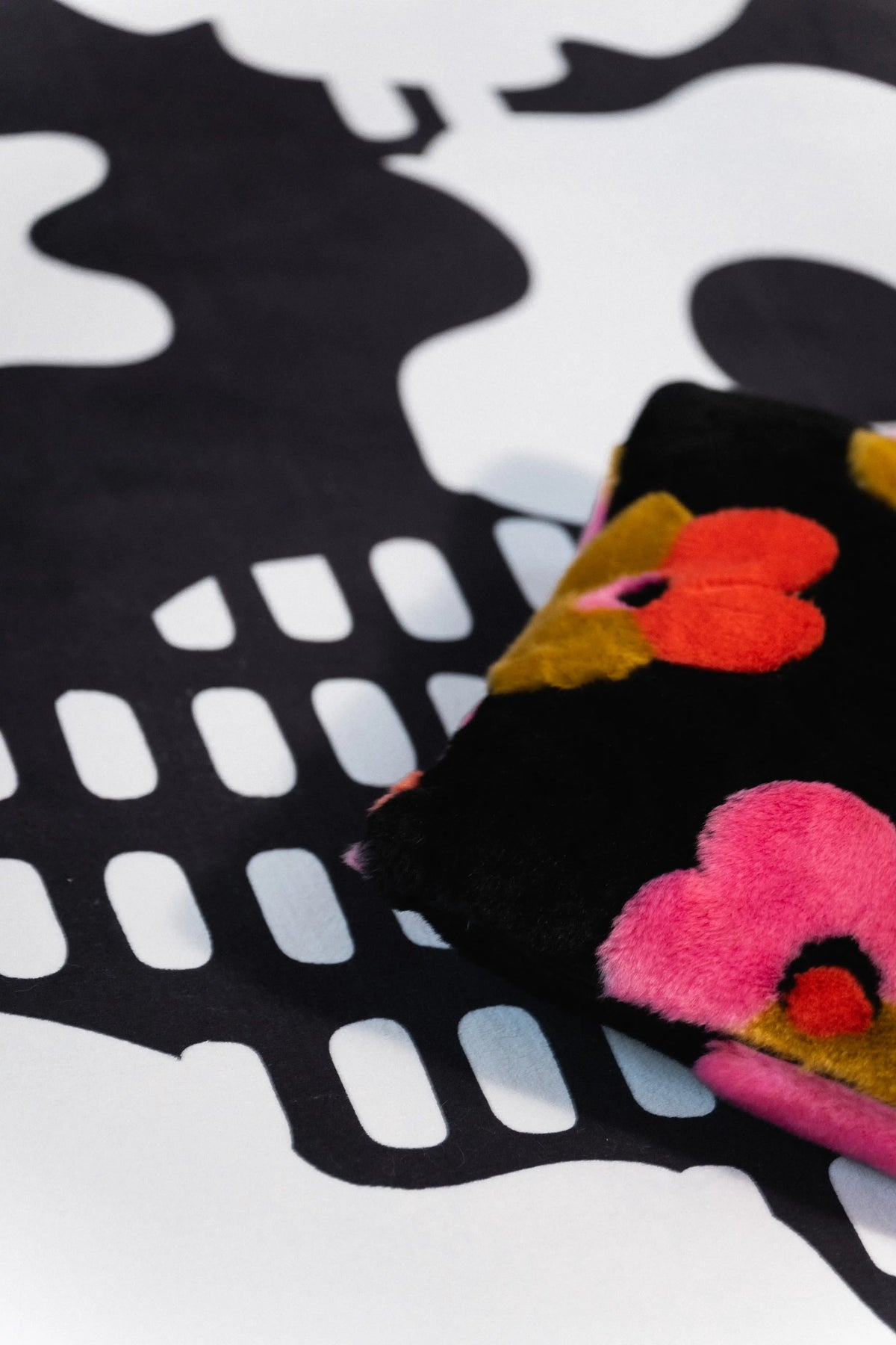 Black pillow with pink, orange, and red flowers resting on a black and white rug with grid marks.