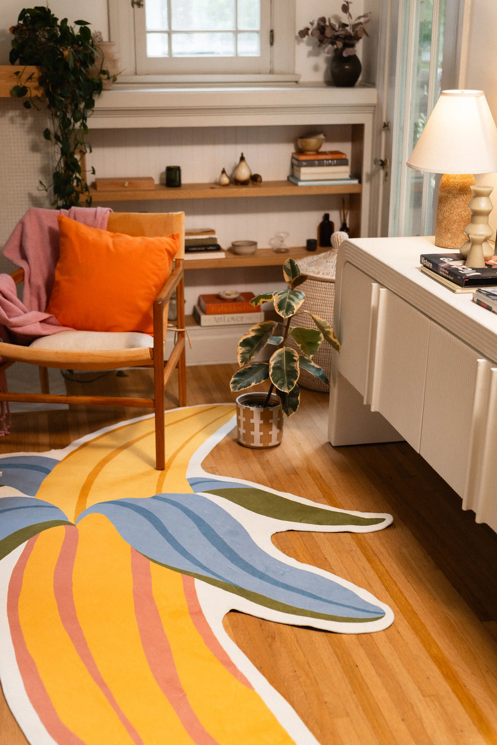 Yellow and blue banana-shaped rug beside a wooden armchair with an orange cushion and white cabinet.