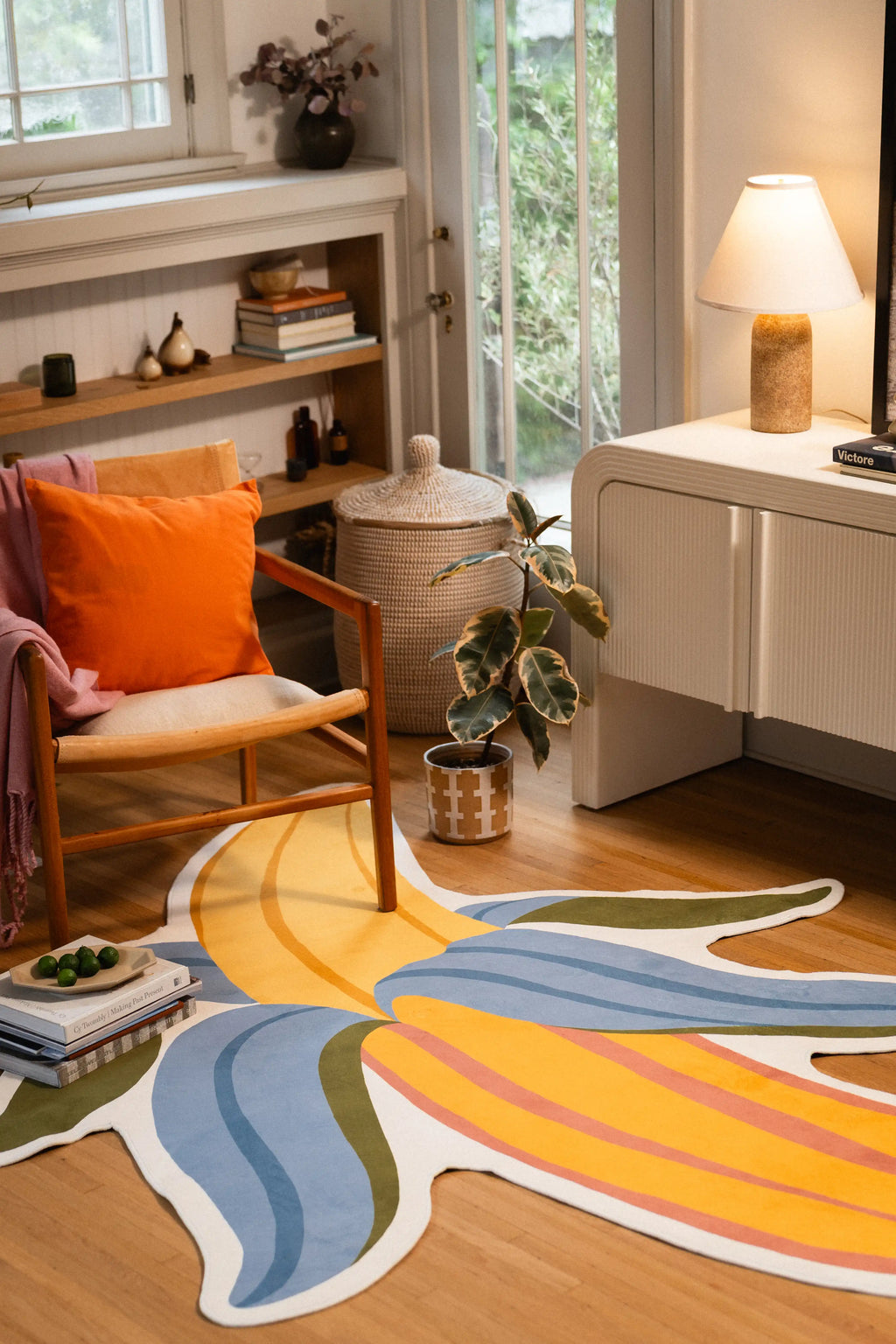 Yellow banana rug with pink stripes and blue and green peel sections beside a wooden chair, potted plant, and white cabinet.