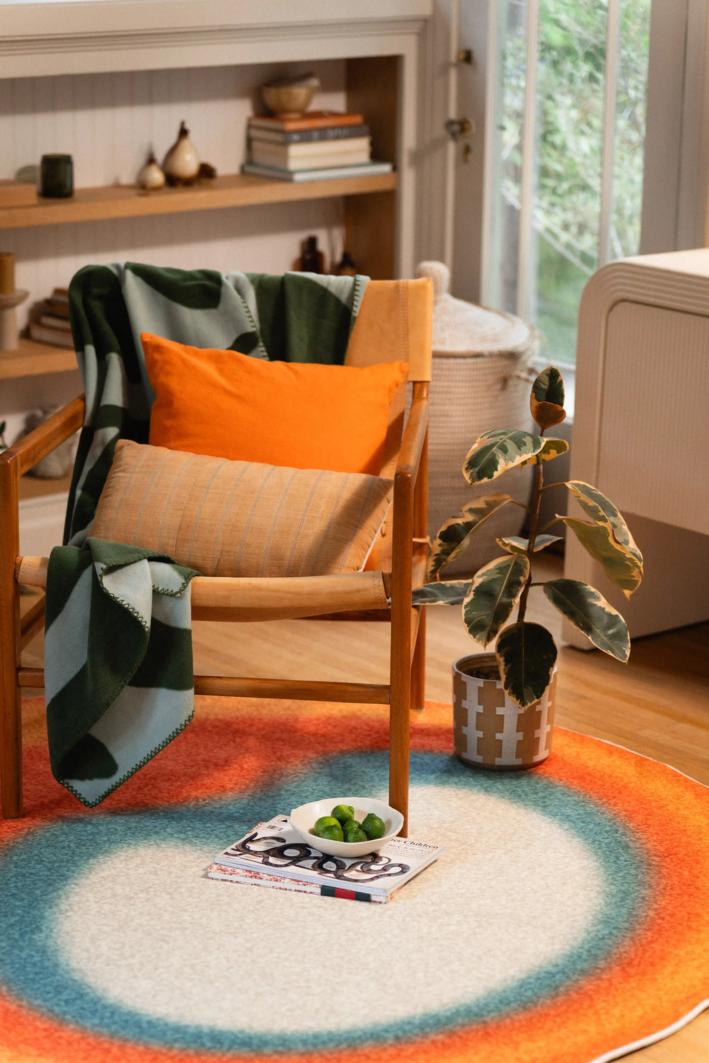 Orange and teal round rug in front of a wooden chair with an orange cushion, beside a potted plant and open magazine.