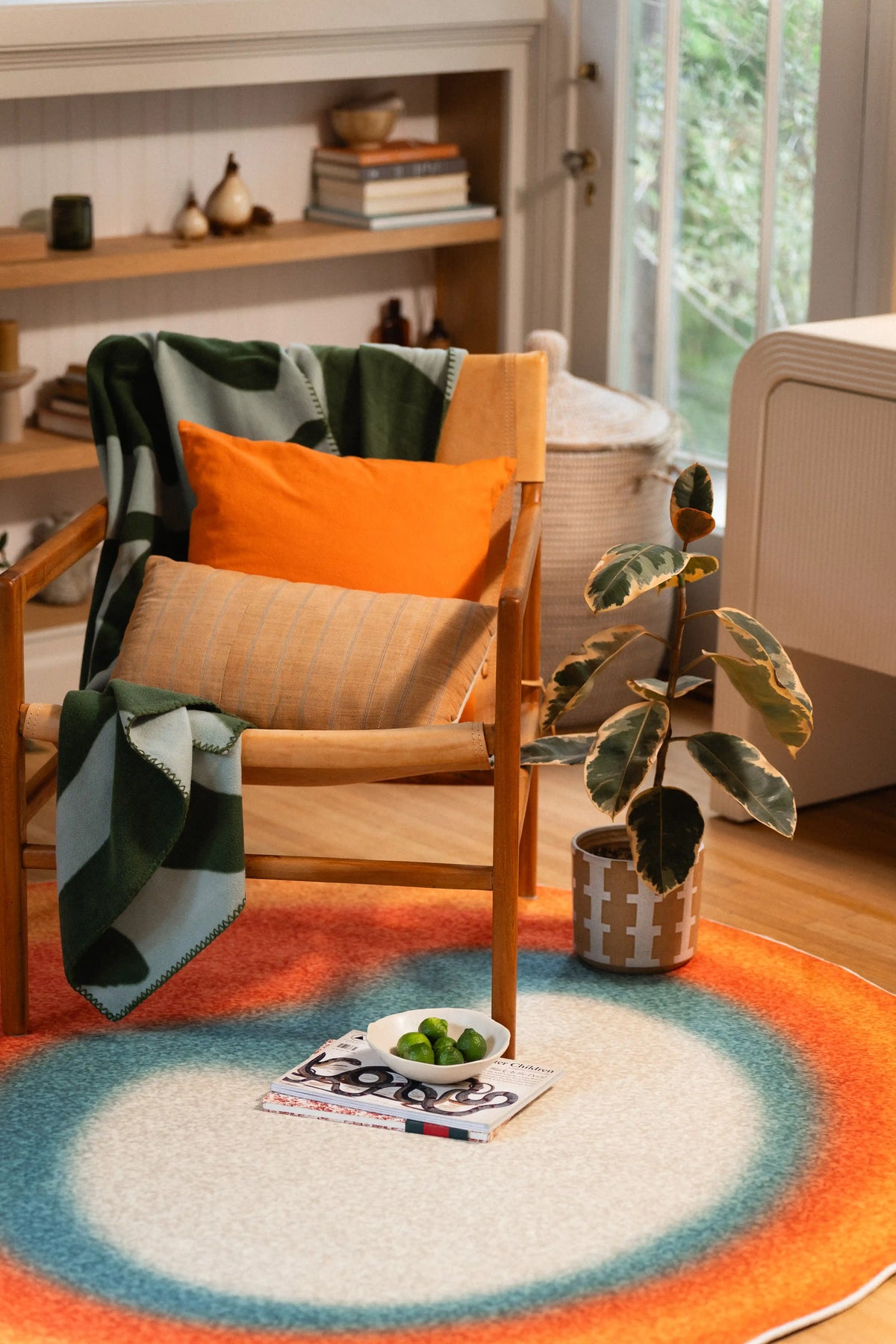 Orange and teal round rug in front of a wooden chair with an orange cushion, beside a potted plant and open magazine.