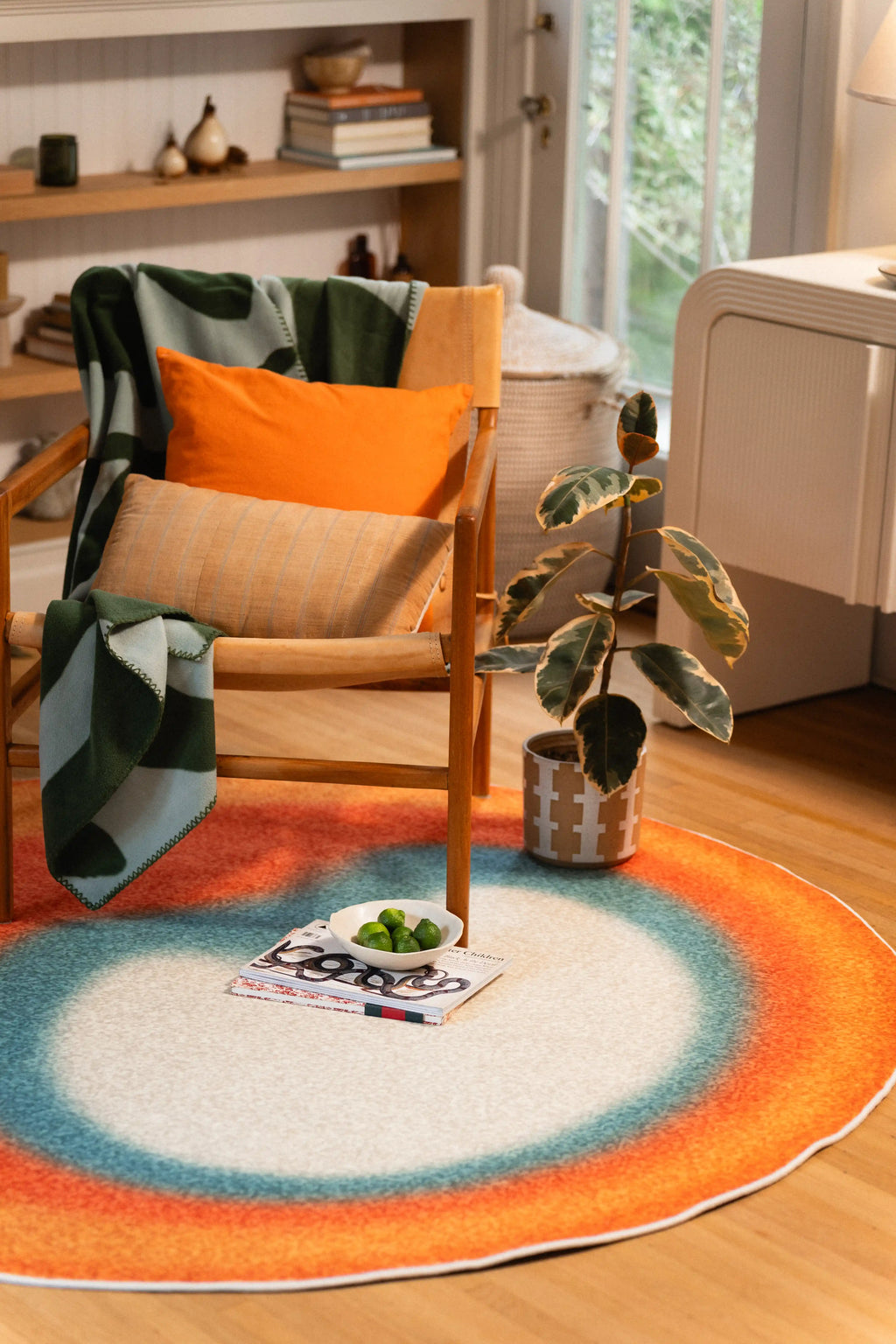 Off-white centered rug with teal and orange rings beneath a wooden armchair, beside a plant and fruit on magazines.