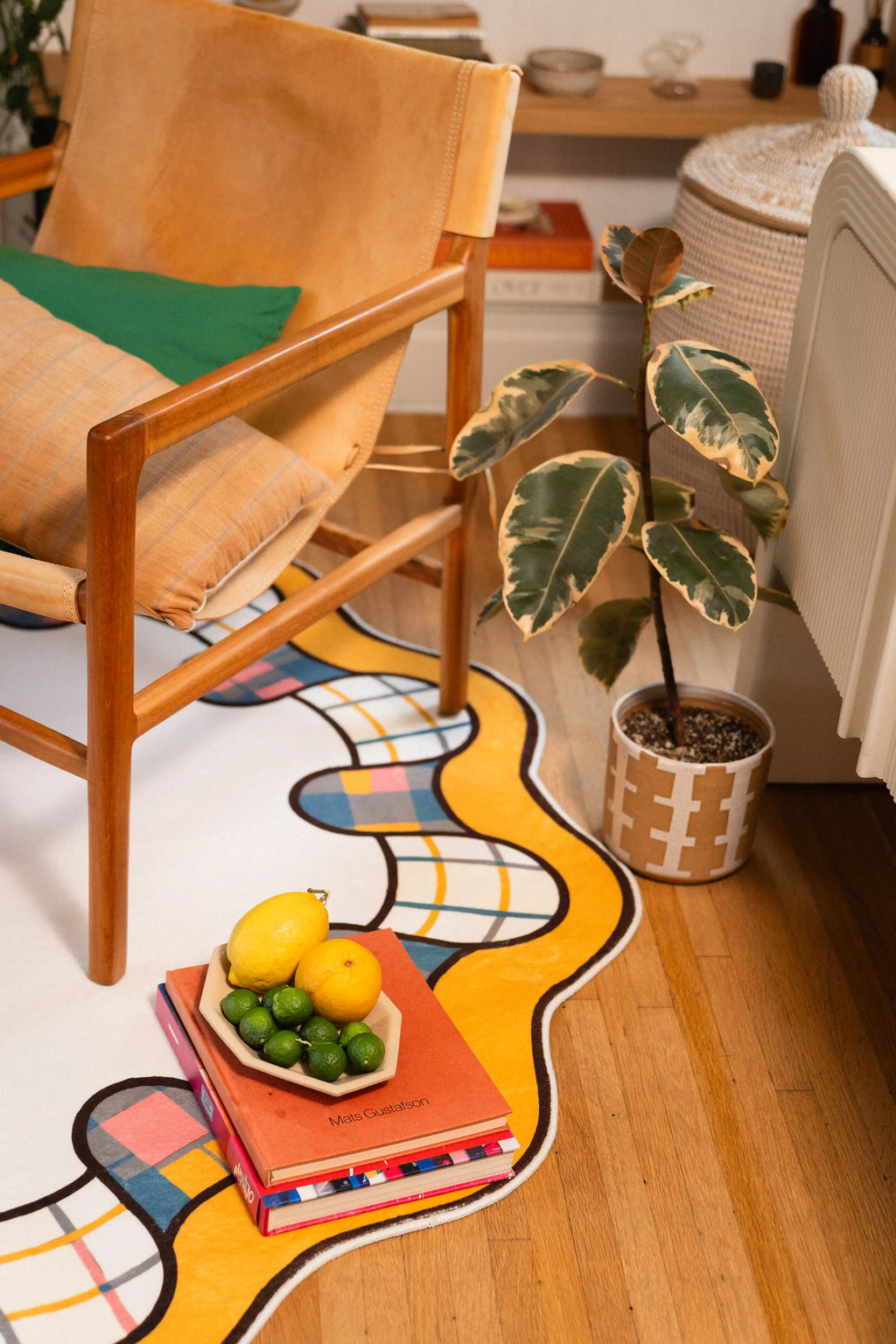 Golden scalloped rug edge with teal, pink, gray, and ivory plaid loops beside a wooden chair and potted plant.