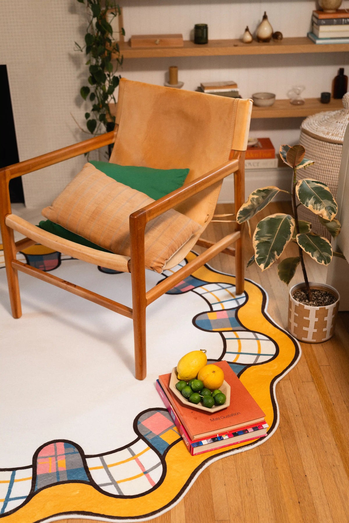 Cream rug with a yellow wavy border beside a wooden armchair, potted plant, and fruit bowl on stacked books.